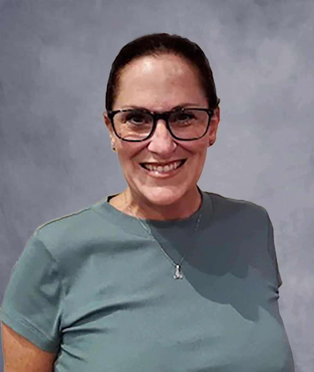 A woman with dark hair, black glasses, and a bright smile, wearing a gray shirt and a necklace, standing in front of a plain beige wall.