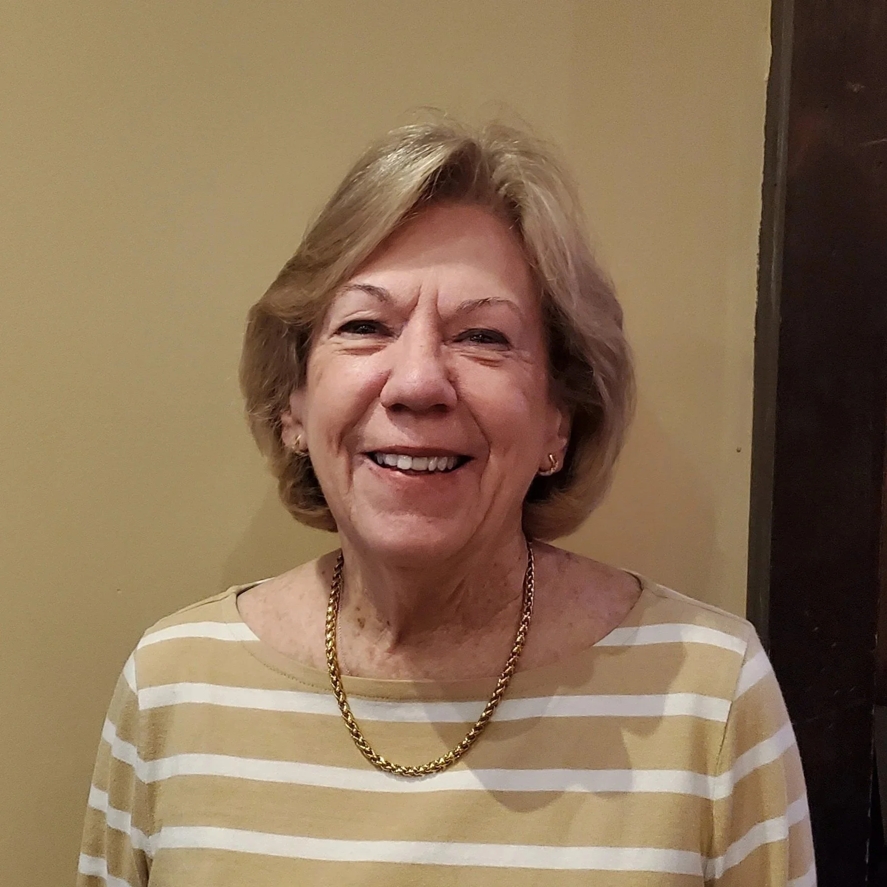 A smiling elderly woman with short, light brown hair, wearing a gold chain necklace and a beige and white striped shirt, standing against a neutral-colored wall.