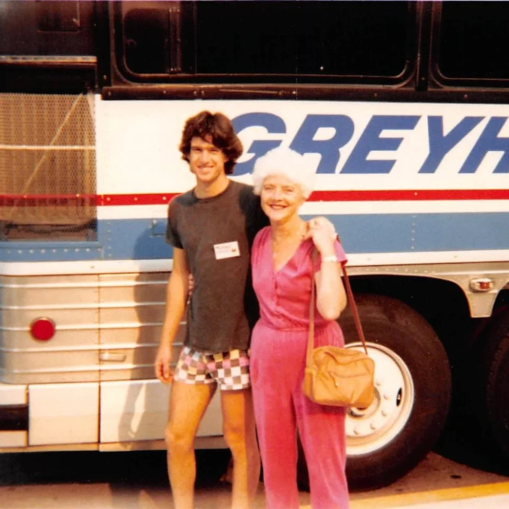 A young man and an elderly woman smiling in front of a Greyhound bus.
