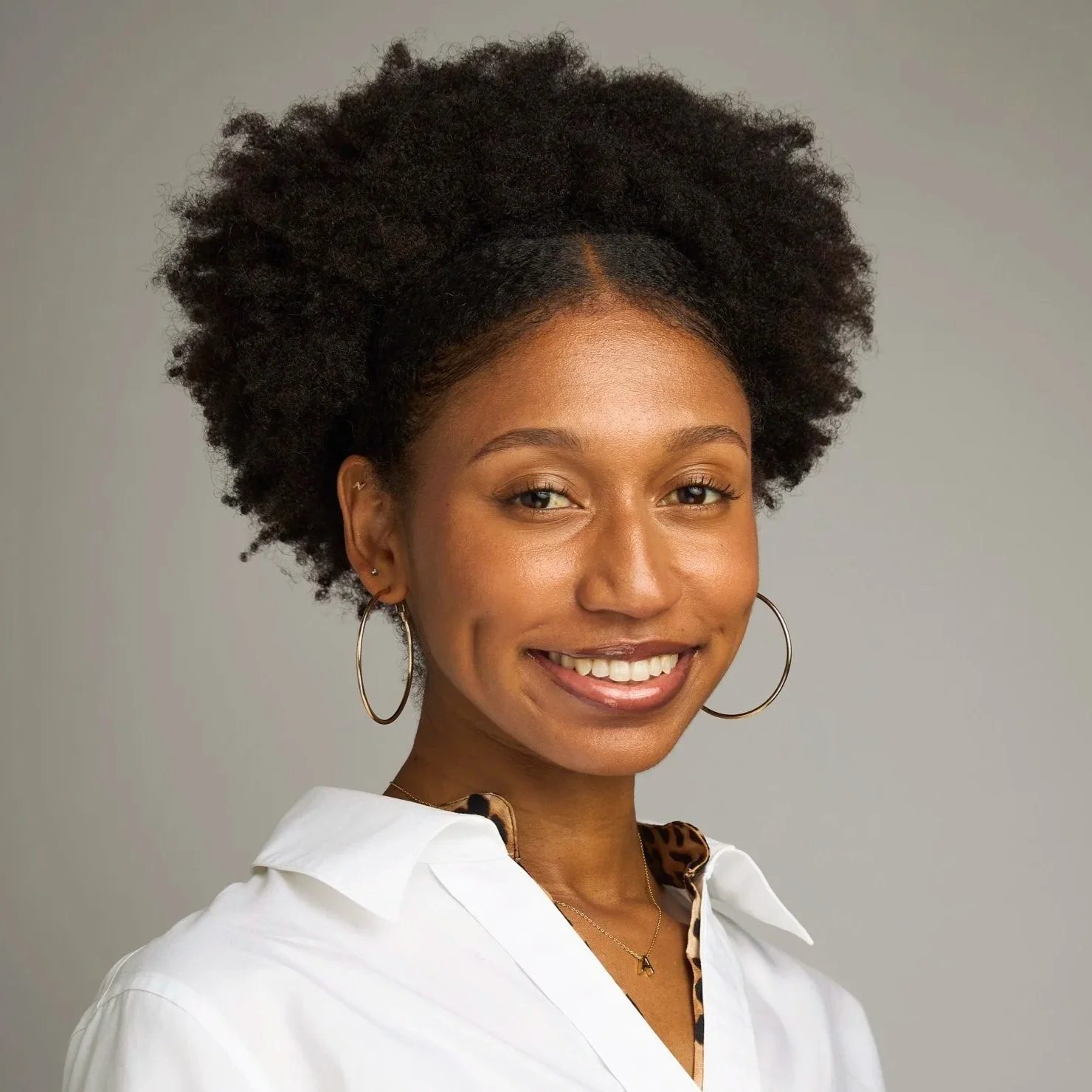 A woman with curly hair wearing a black headband and a yellow blouse with small black stars, smiling against a beige wall.