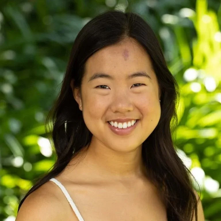 A young woman with long dark hair smiling outdoors with green foliage in the background.