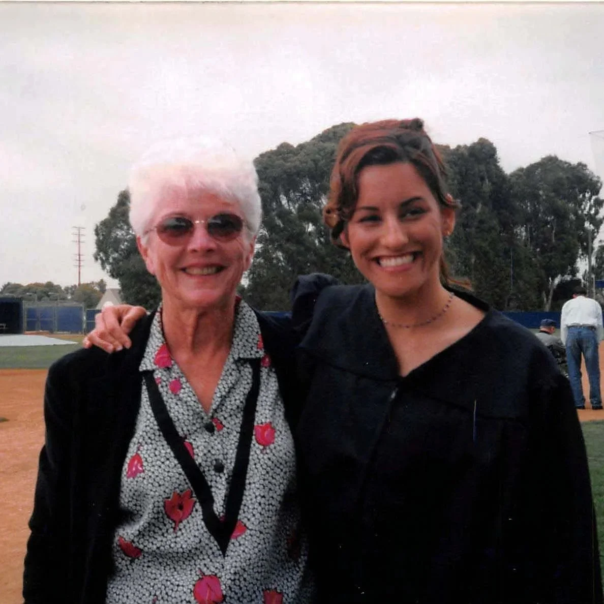 Two women standing on a baseball field, smiling and posing for the photo. One woman has short white hair and glasses, wearing a patterned blouse with pink flowers, and a black jacket. The other woman has shoulder-length brown hair, wearing a black coat and a choker necklace. There are trees and a few other people in the background.