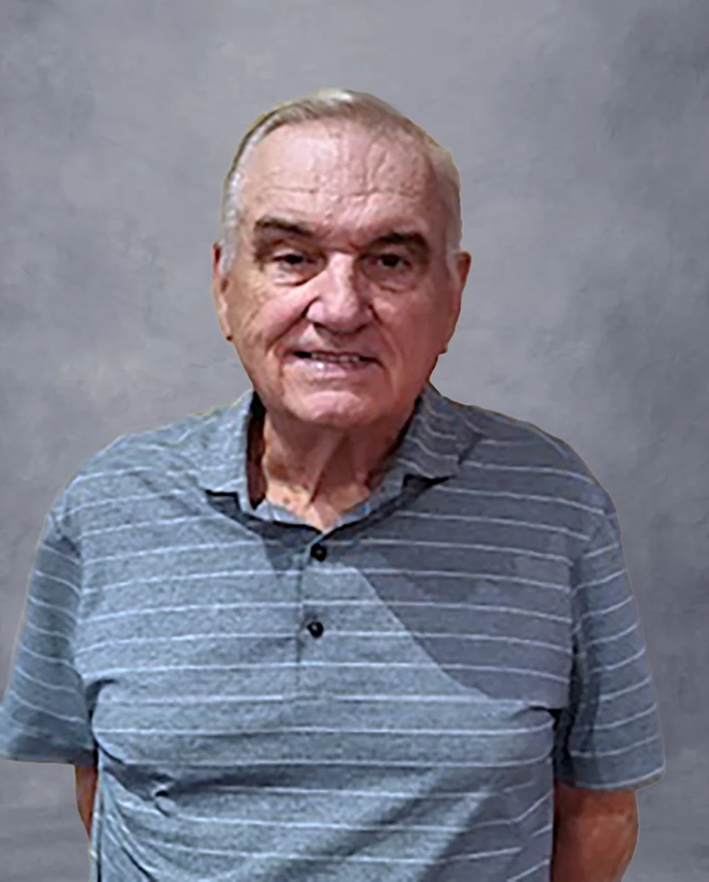 An elderly man with gray hair smiling, wearing a gray striped collared shirt, standing against a light-colored wall.