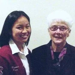 A young woman and an elderly woman standing together, smiling for the photo. The young woman is wearing a burgundy uniform with a white collar, and the elderly woman has short white hair and is wearing glasses and a dark jacket.