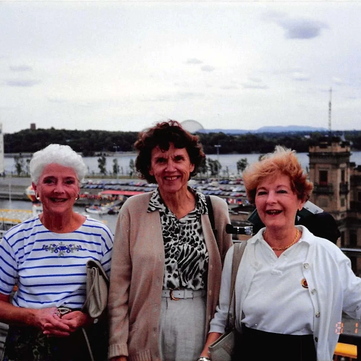 Three women standing together outdoors, with a river and a cityscape in the background, smiling at the camera.