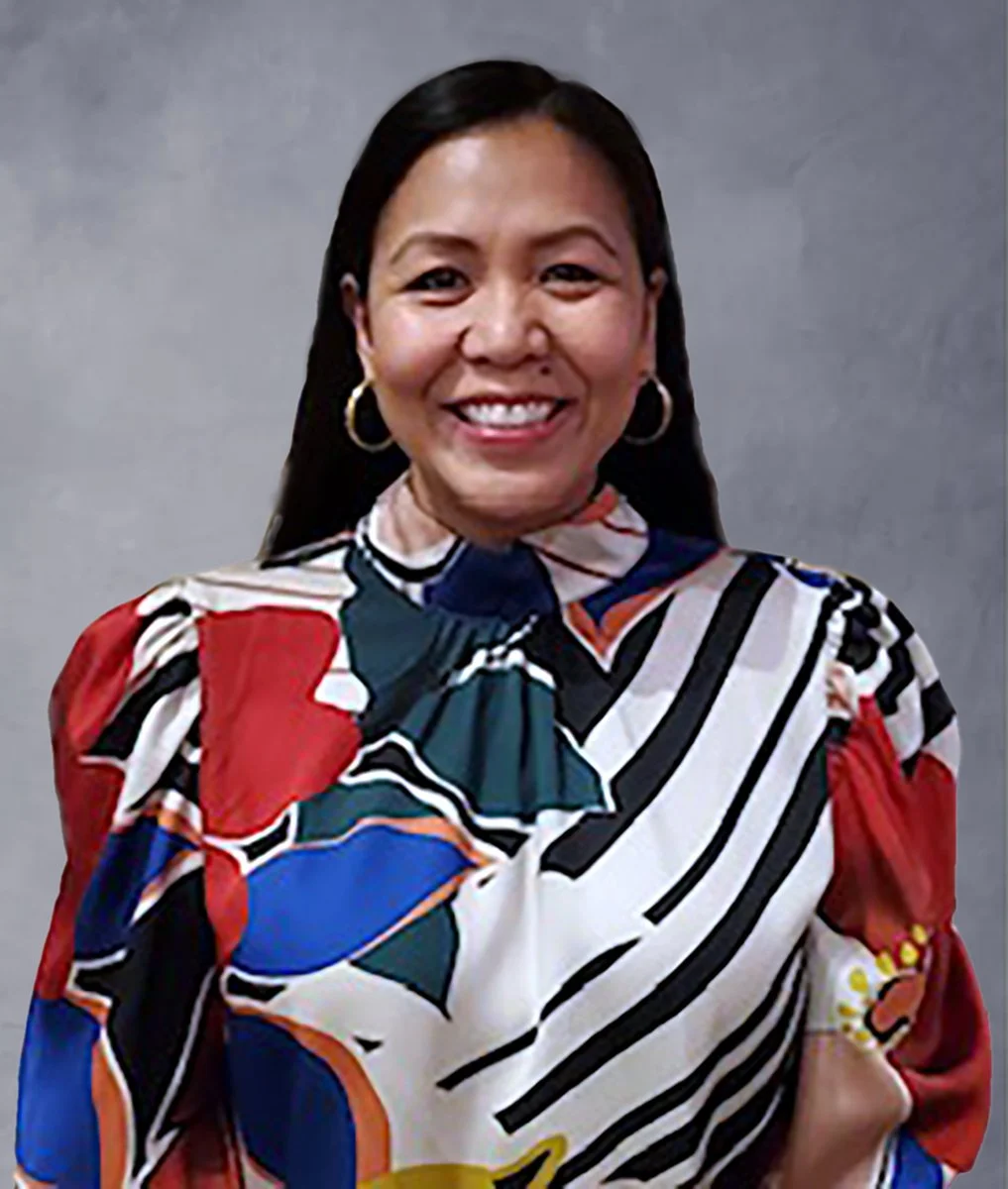 A woman smiling, wearing a colorful patterned blouse with red, black, white, blue, and green, and gold hoop earrings, standing against a plain beige wall.