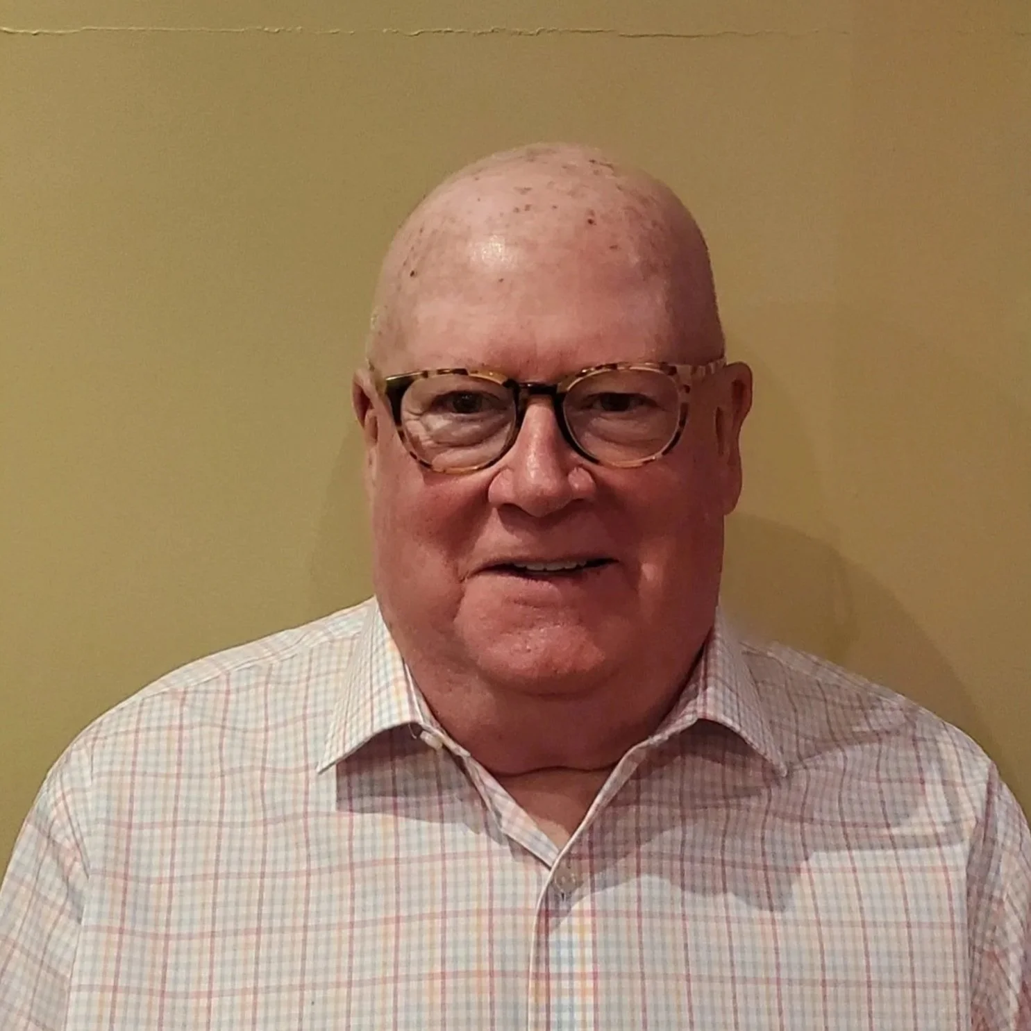 A man with glasses, short light hair, and a light checkered shirt standing against a beige wall.