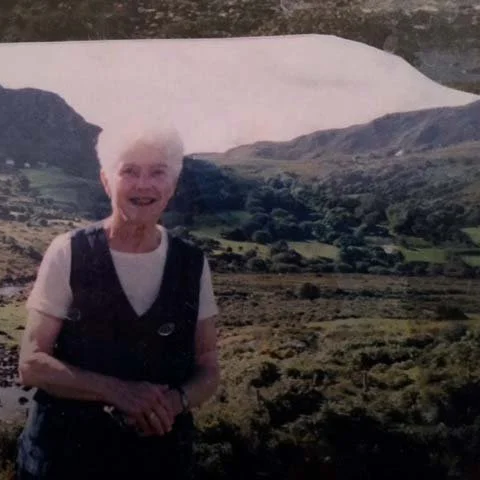 Older woman with short white hair standing outdoors with a scenic mountainous valley in the background.