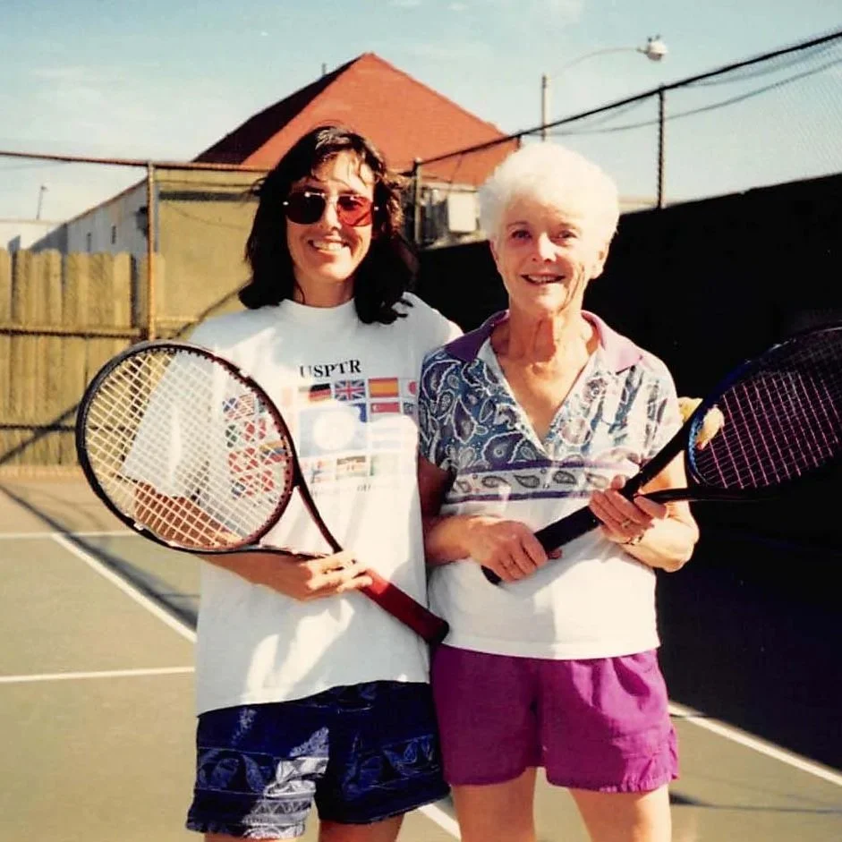 Two women stand on a tennis court holding tennis rackets, smiling, with a building, a fence, and blue sky in the background.