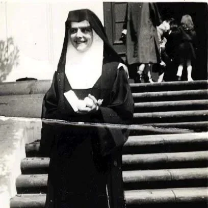 A woman dressed as a nun smiling and holding a book, standing in front of steps with other people in the background.