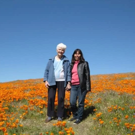 Two women standing hand in hand in a field of orange flowers under a clear blue sky.