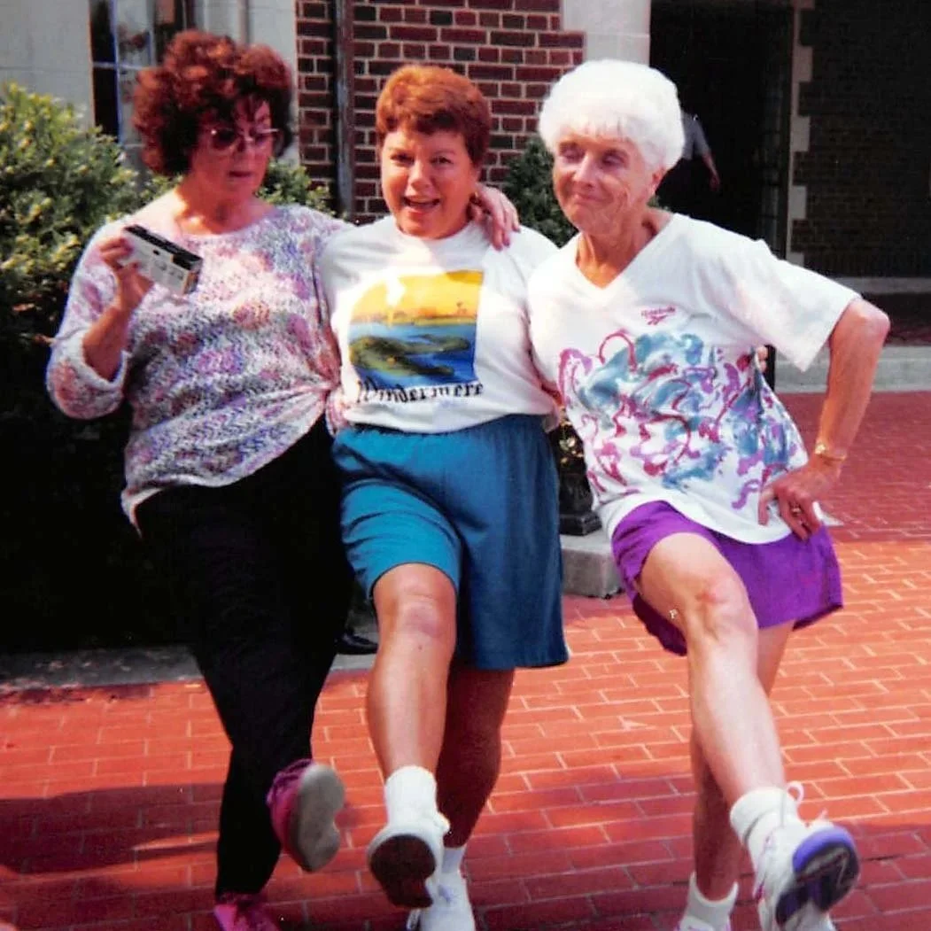 Three women standing outdoors in front of a brick building, lifting their legs in a playful manner. The woman on the left has curly red hair, glasses, and is holding a camera. The woman in the middle has short brown hair and is smiling. The woman on the right has white hair, is wearing a white t-shirt with colorful designs and purple shorts.