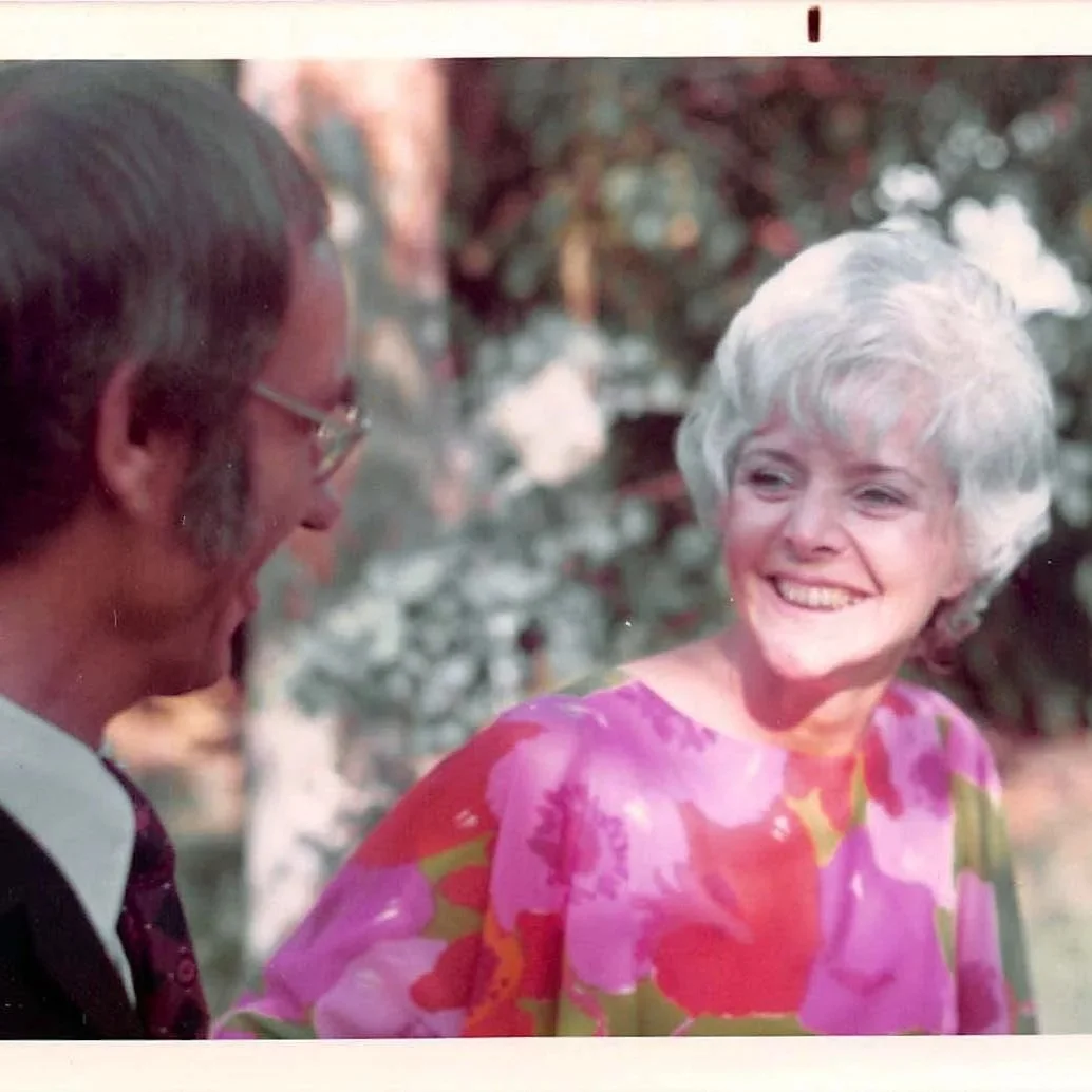 A woman with short gray hair smiling at a man with glasses, dark hair, and brown facial hair, outdoors with trees in the background.