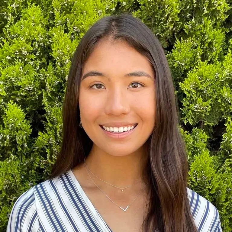 A young woman with long dark hair smiling in front of green bushes, wearing a striped top and layered necklaces.