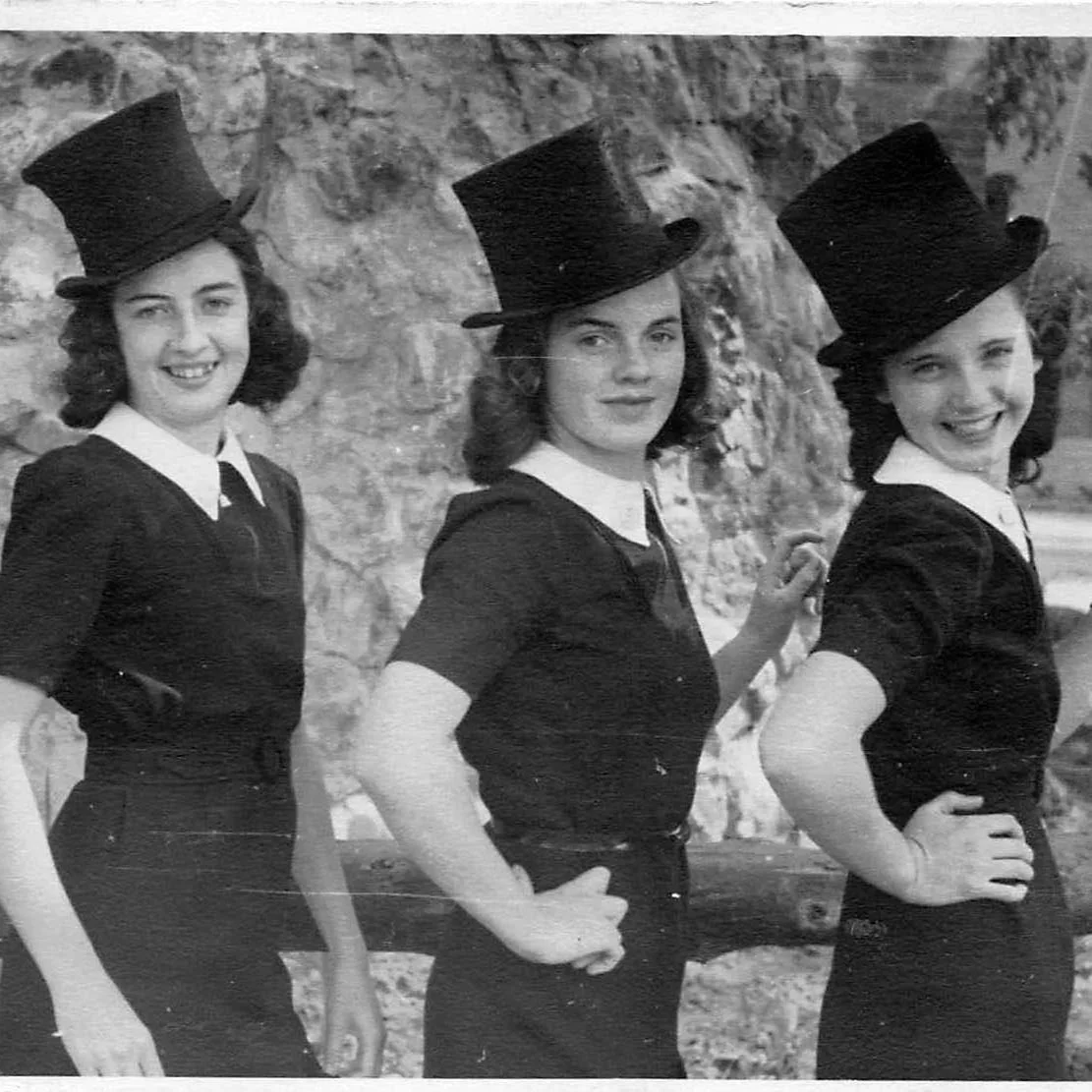 Three young women in old-fashioned dark dresses with white collars, wearing tall hats, standing outdoors in front of a stone wall and a tree, smiling and posing for the photograph.