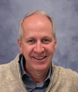 Smiling middle-aged man with short light-colored hair, wearing a dark patterned shirt, in front of a beige wall.