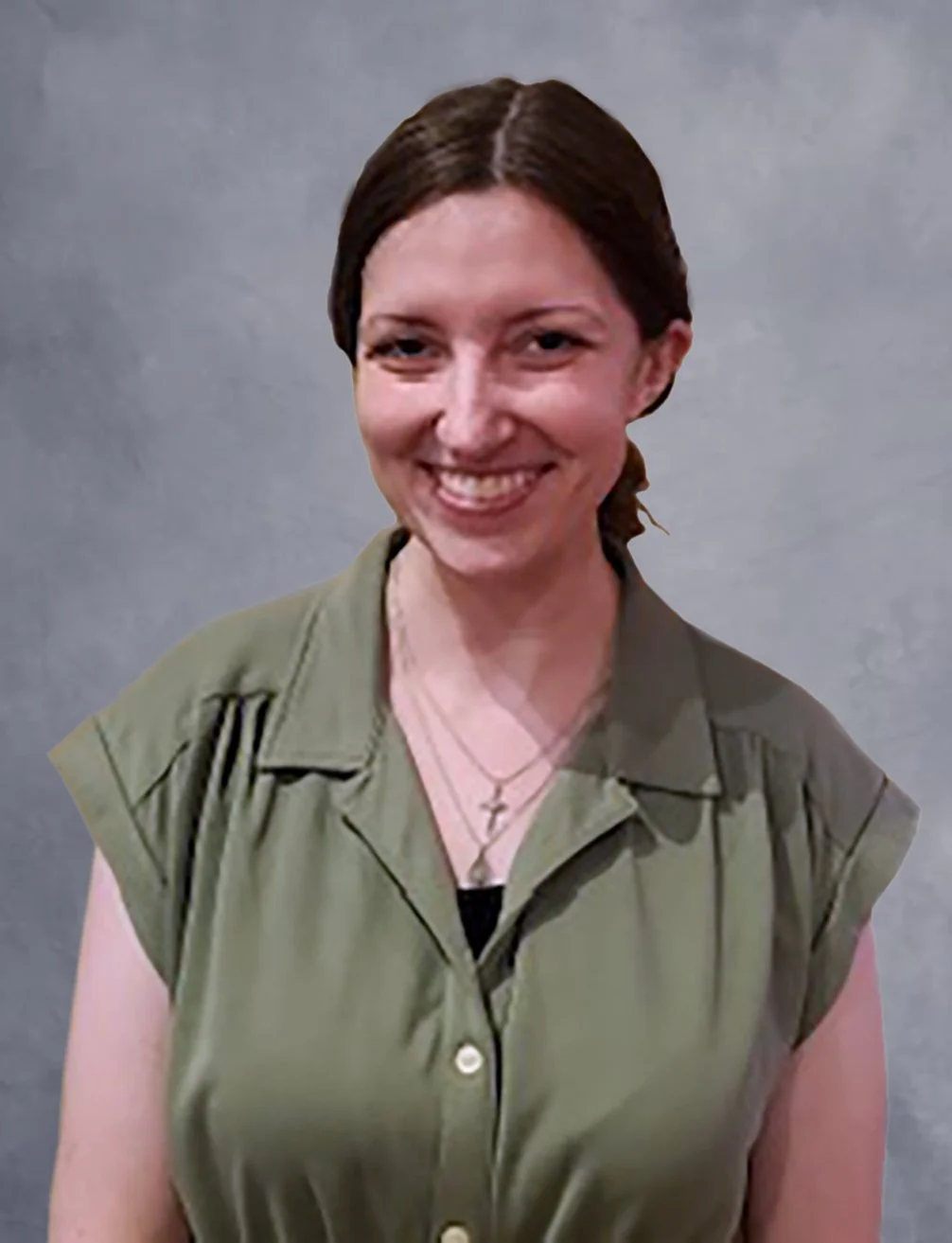 A smiling woman with dark brown hair pulled back, wearing an olive-green shirt and layered necklaces with cross pendants, standing against a beige-colored wall.