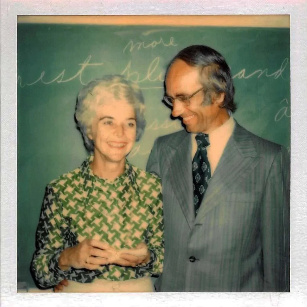 A woman and a man smiling and looking at each other, standing in front of a green chalkboard with handwritten notes.