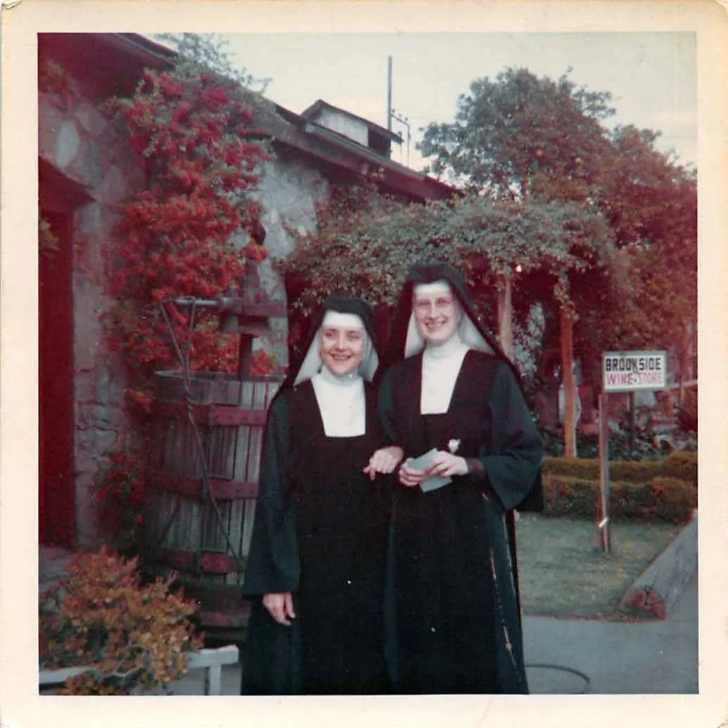 Two women dressed as nuns standing outdoors near a stone wall, plants, and a sign that reads 'Brookside WINE STORE'.