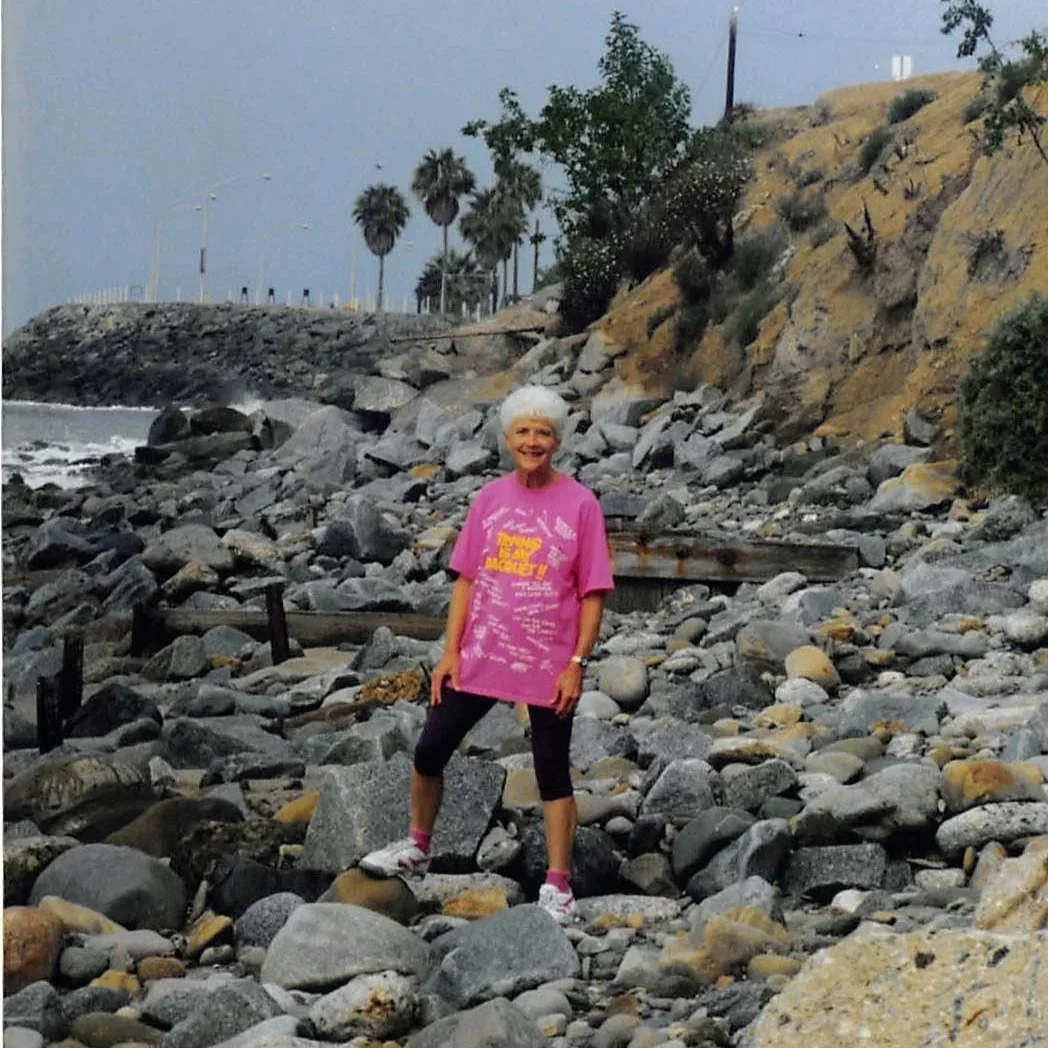 An older woman with white hair stands on a rocky beach, smiling at the camera, wearing a bright pink T-shirt, black leggings, and white sneakers with pink accents, with the ocean, rocks, and hillside with trees in the background.