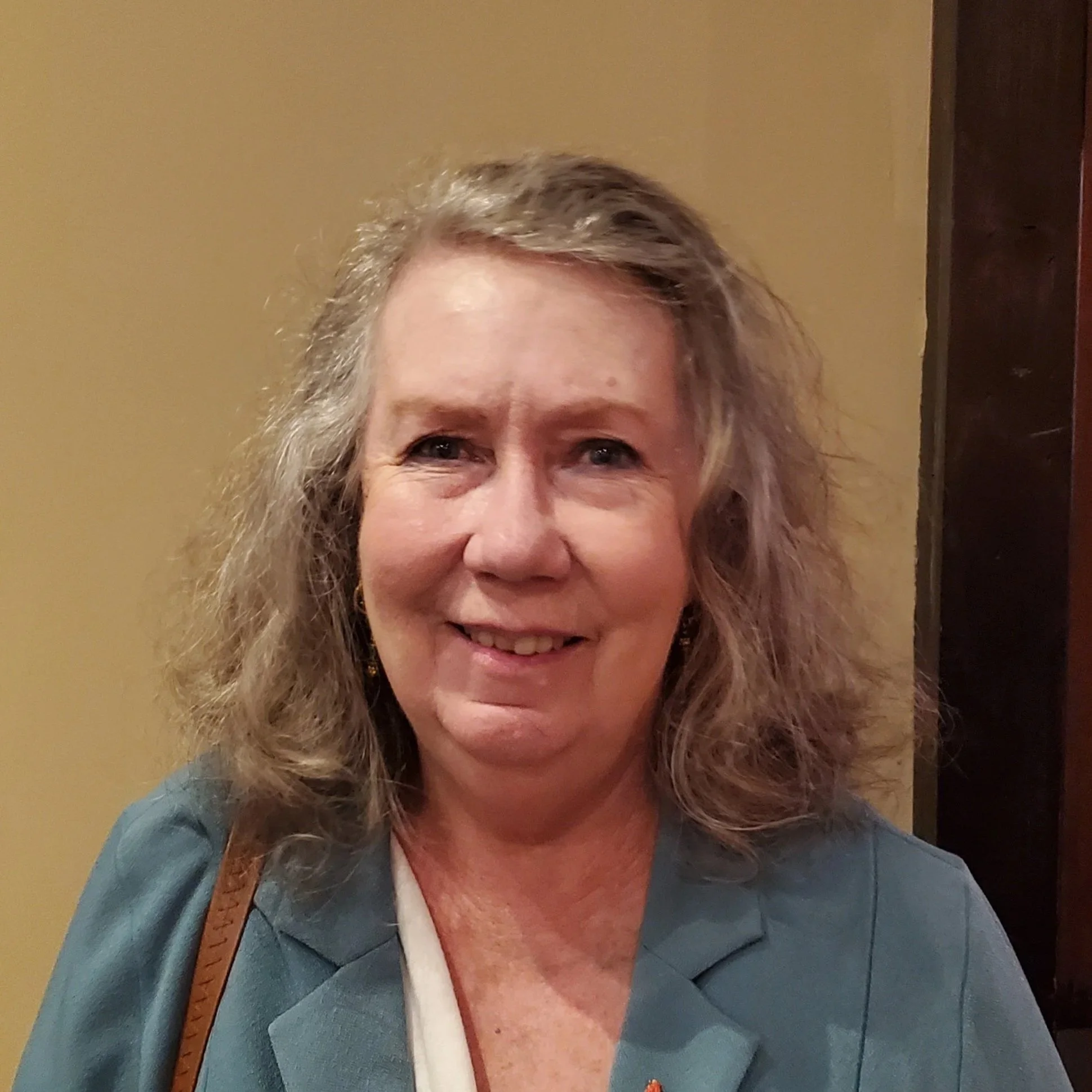 A smiling middle-aged woman with shoulder-length, wavy, light brown hair, wearing a teal blazer, a white top, and a brown strap from a bag or purse, standing indoors against a beige wall next to a wooden door or panel.