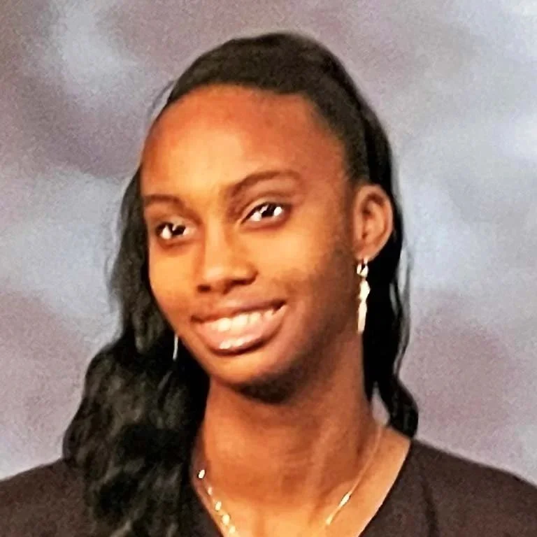 A young woman with long, wavy black hair, wearing earrings and a necklace, smiling in front of a plain background.