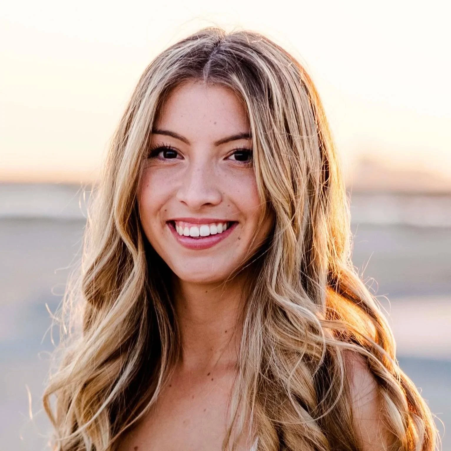 A young woman smiling at the beach during sunset with long wavy blonde hair.