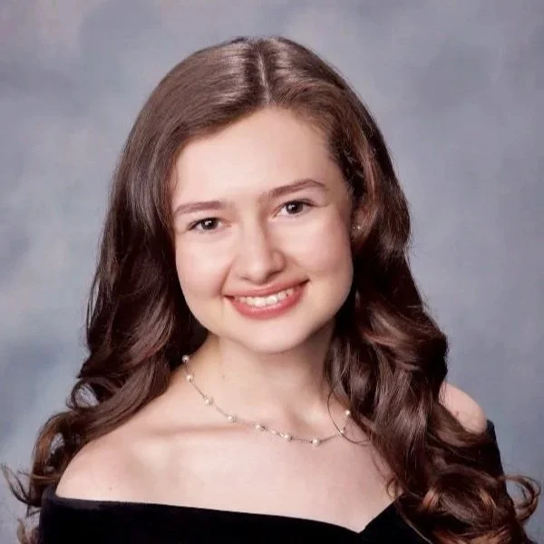 A young woman with long, curly brown hair and a bright smile, wearing a black off-shoulder top and a delicate necklace in front of a plain gray background.