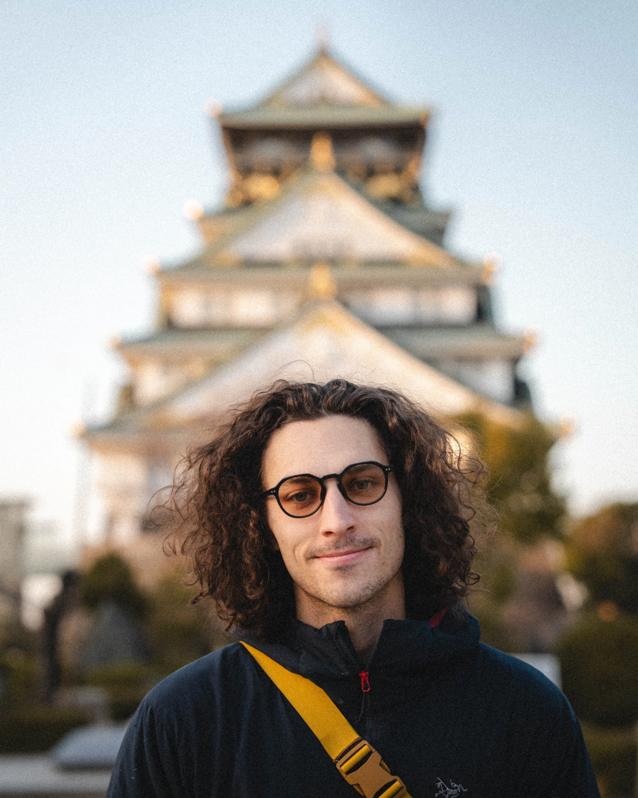 A young man with curly brown hair, wearing glasses and a black jacket, standing outdoors with a blurred traditional multi-tiered building in the background.