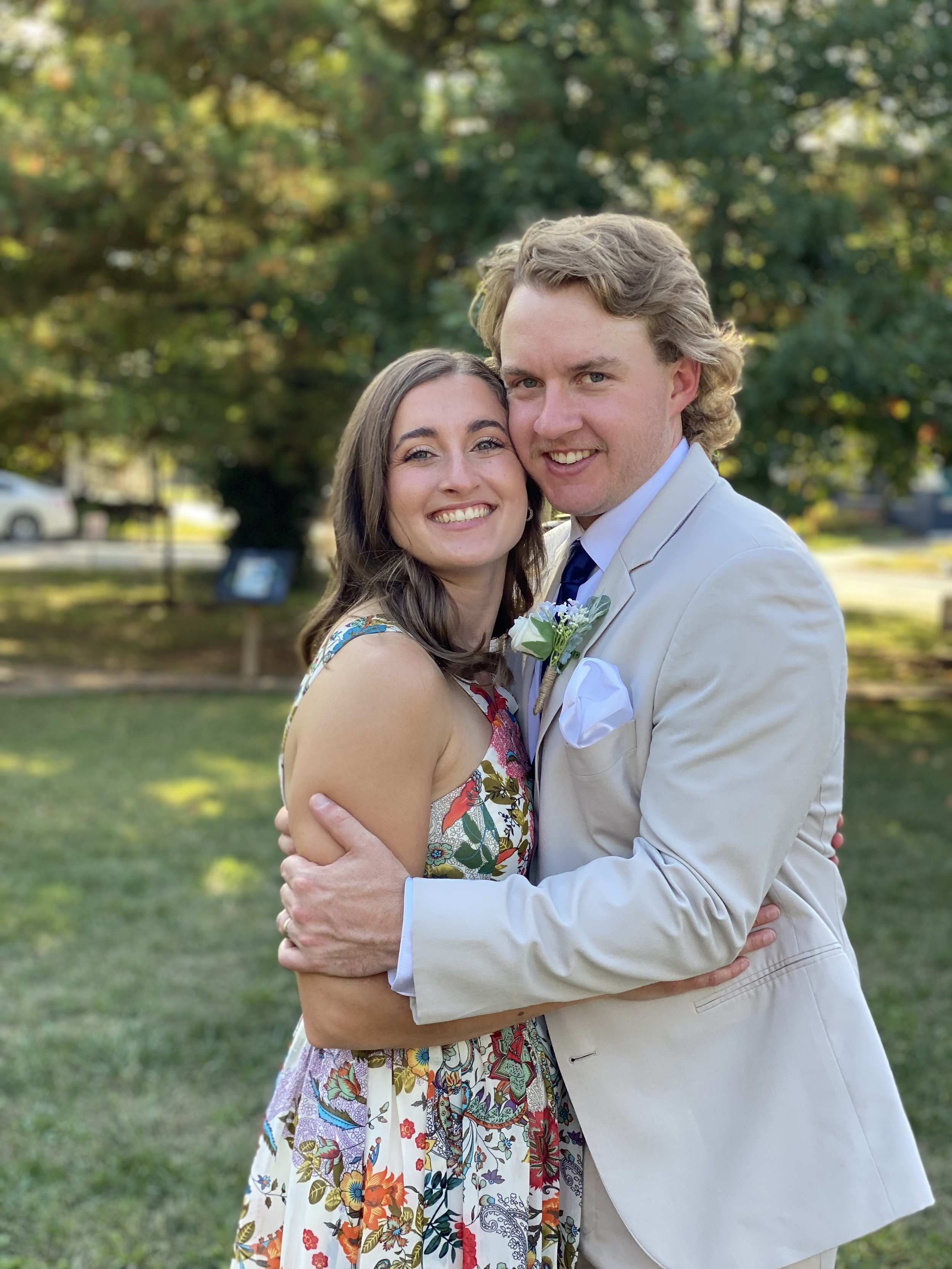 A smiling couple hugging outdoors in a park, with trees and cars in the background. The woman has long brunette hair and is wearing a colorful, floral dress. The man has blonde hair, is dressed in a light beige suit with a white shirt and a dark tie, and has a white pocket square and boutonnière.