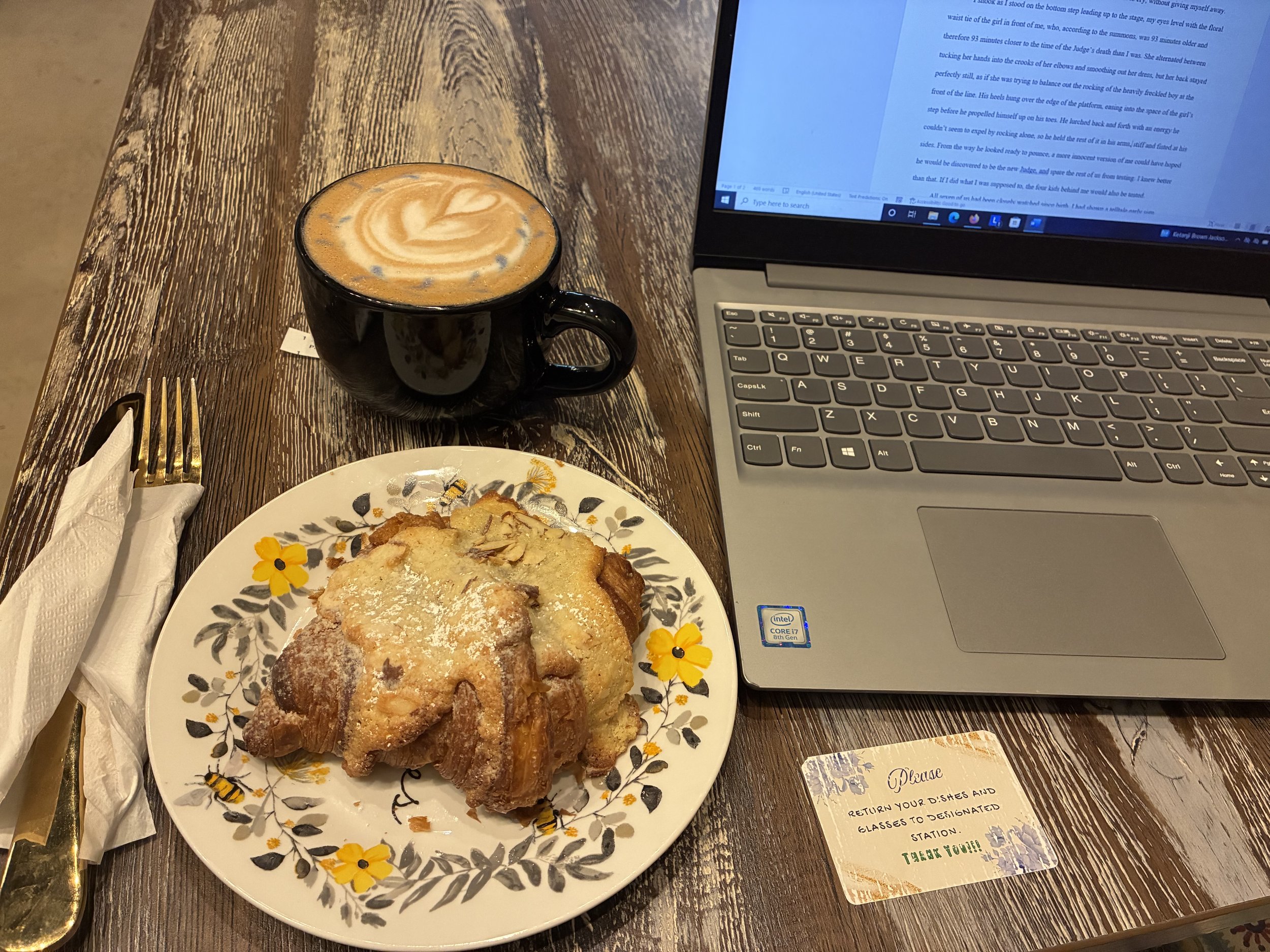A table with a black coffee mug, a plate with a pastry, a fork and napkin, and a laptop. The pastry appears to be a cinnamon roll or similar baked good, dusted with powdered sugar. The laptop is open with blue text on the screen. The table is wooden.