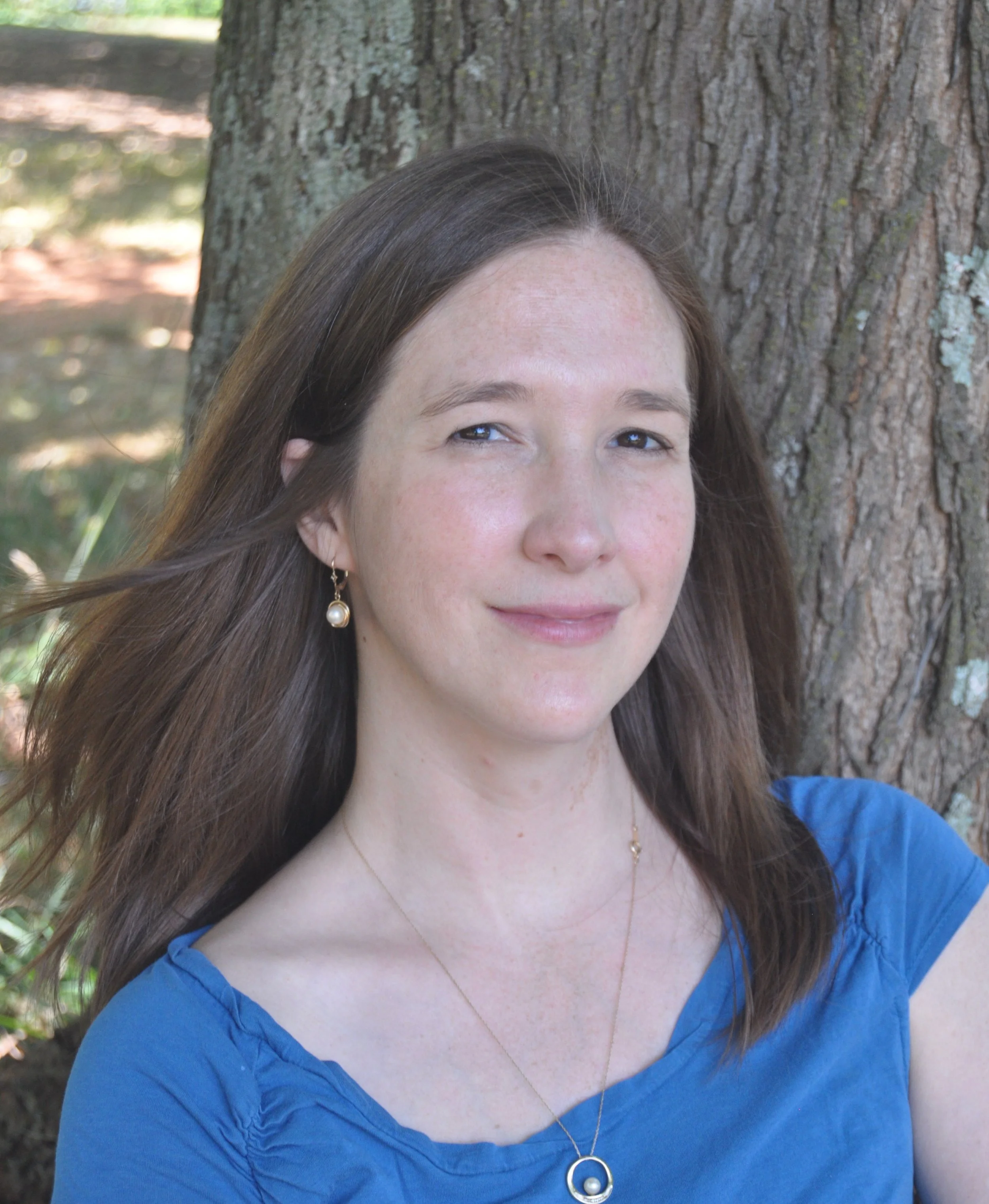 A woman with brown hair wearing a blue top and gold jewelry standing outdoors in front of a large tree trunk.