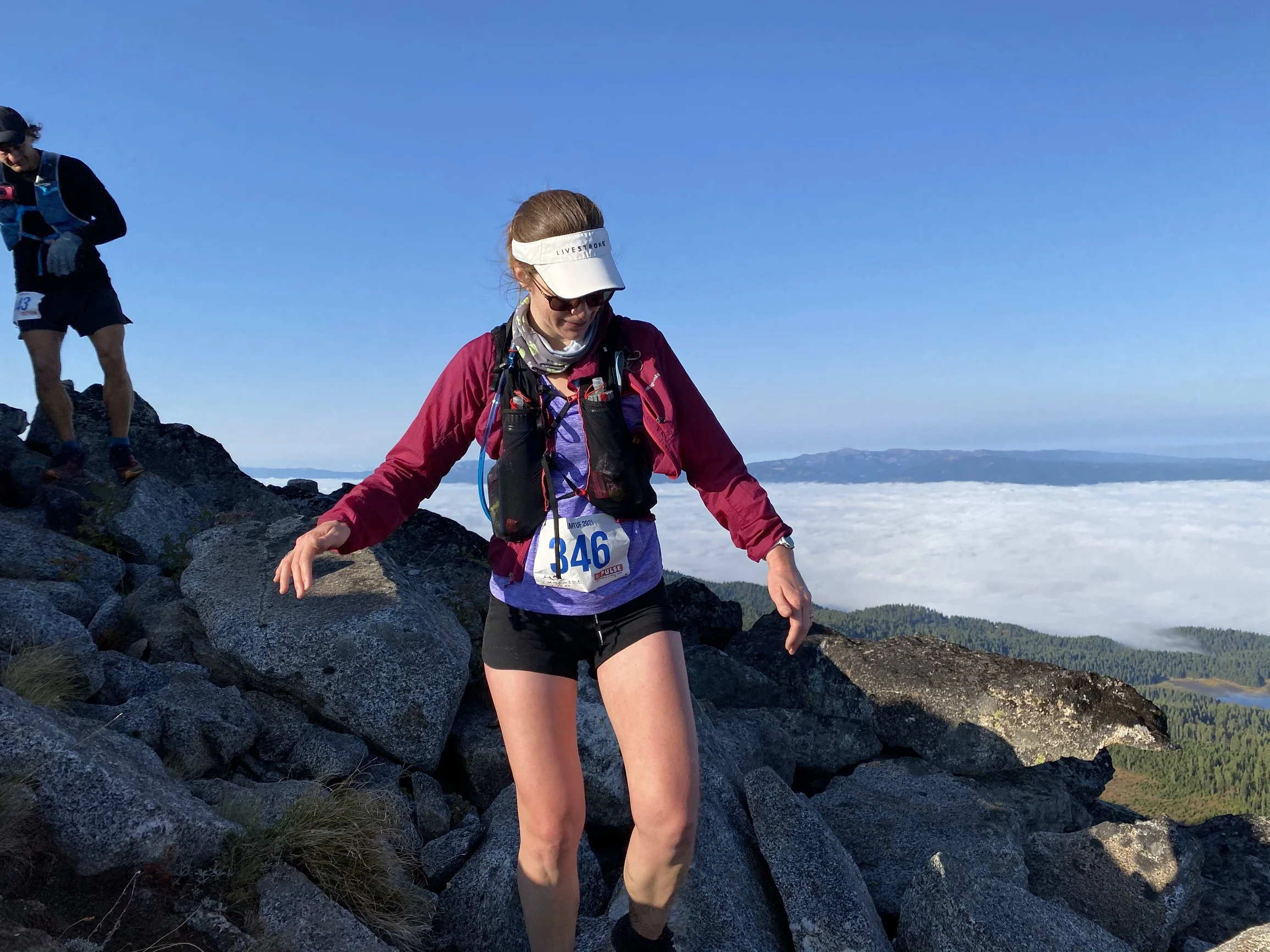 A woman trail running on rocky terrain on a mountain, with a cloudy valley below and a clear blue sky.