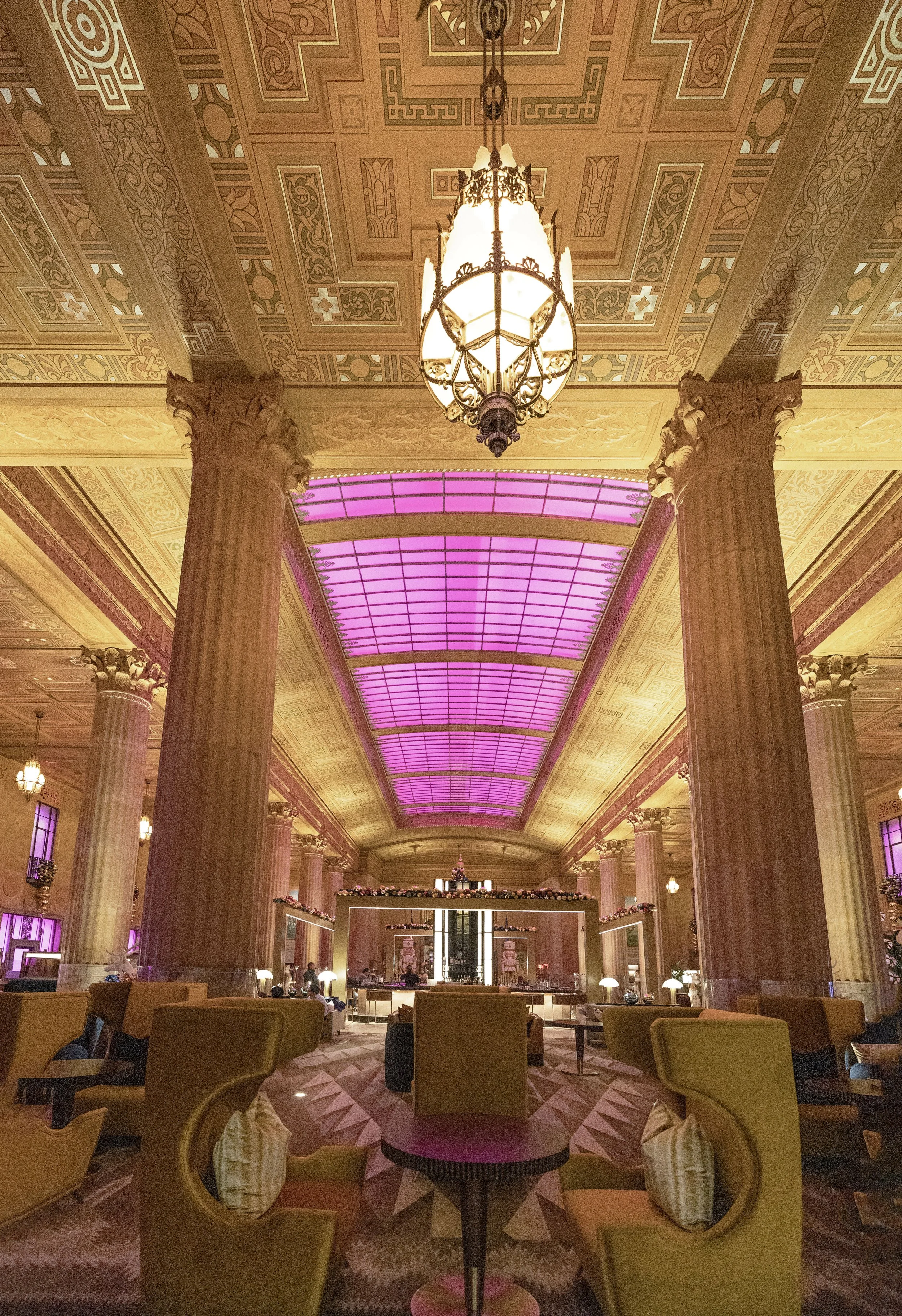 Elegant hotel lobby with tall columns, ornate ceiling, stained glass upper walls, and a pink-lit skylight.  The National Hotel, Oklahoma City.