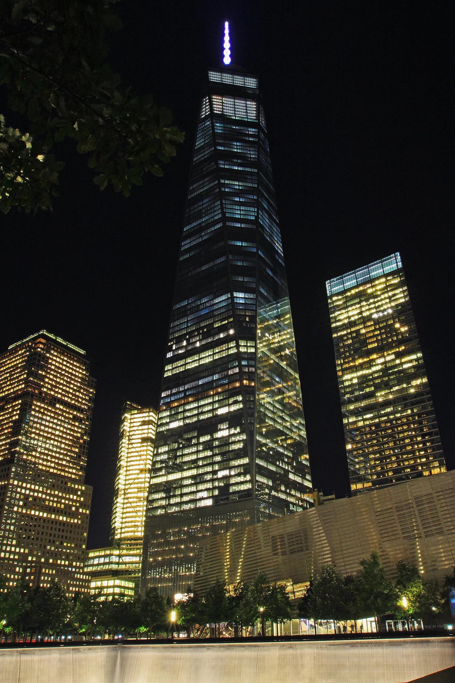 Night view of a tall, illuminated skyscraper with a glowing light on top, surrounded by other high-rise buildings with lit windows, trees, and a low wall at the base. Freedom Tower New York City.