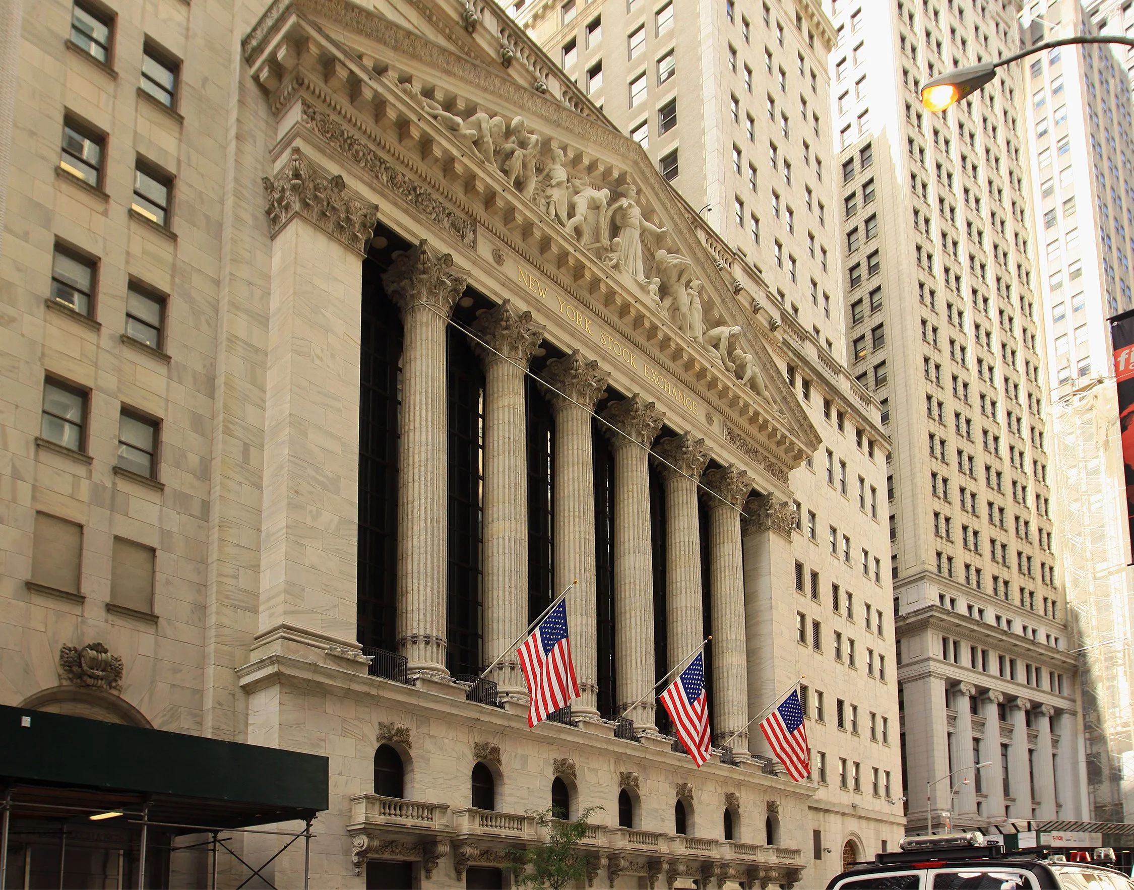 Front view of the New York Stock Exchange building with five American flags displayed outside. Classical architecture with large columns and detailed sculptures on the facade.