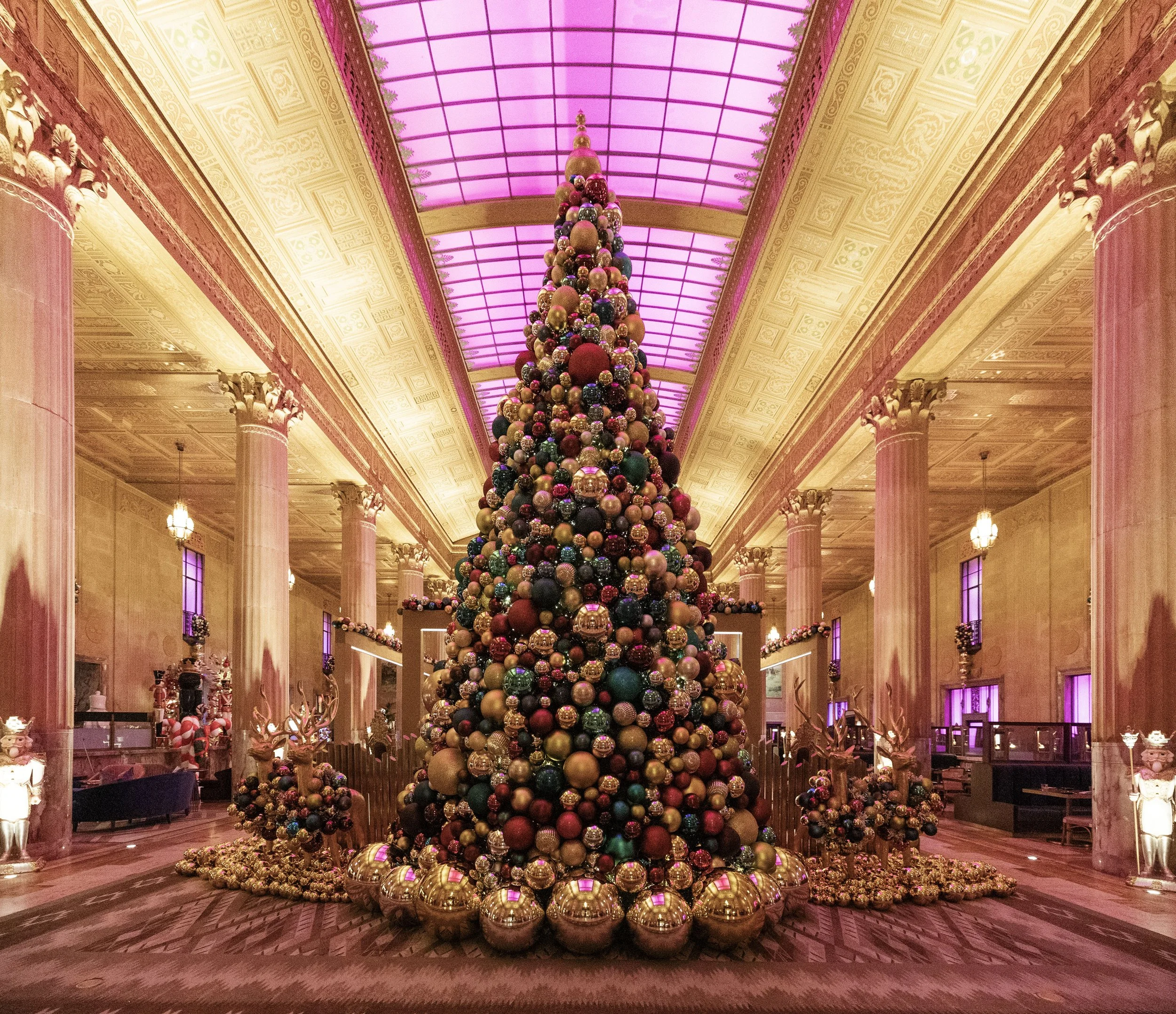A large Christmas tree decorated with multicolored ornaments, inside an elegant, brightly lit hall with pink and purple lighting, tall columns, and ornate ceiling details. The National Hotel, Oklahoma City.