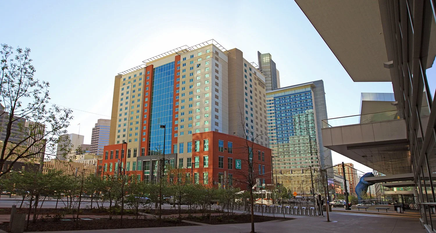 Denver Embassy Suites next to the Denver Convention Center, trees, and a sidewalk with seating and benches.