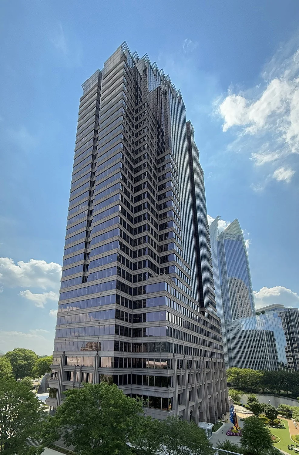 A tall modern skyscraper with glass windows reflecting the sky, surrounded by other high-rise buildings and greenery on a sunny day.
