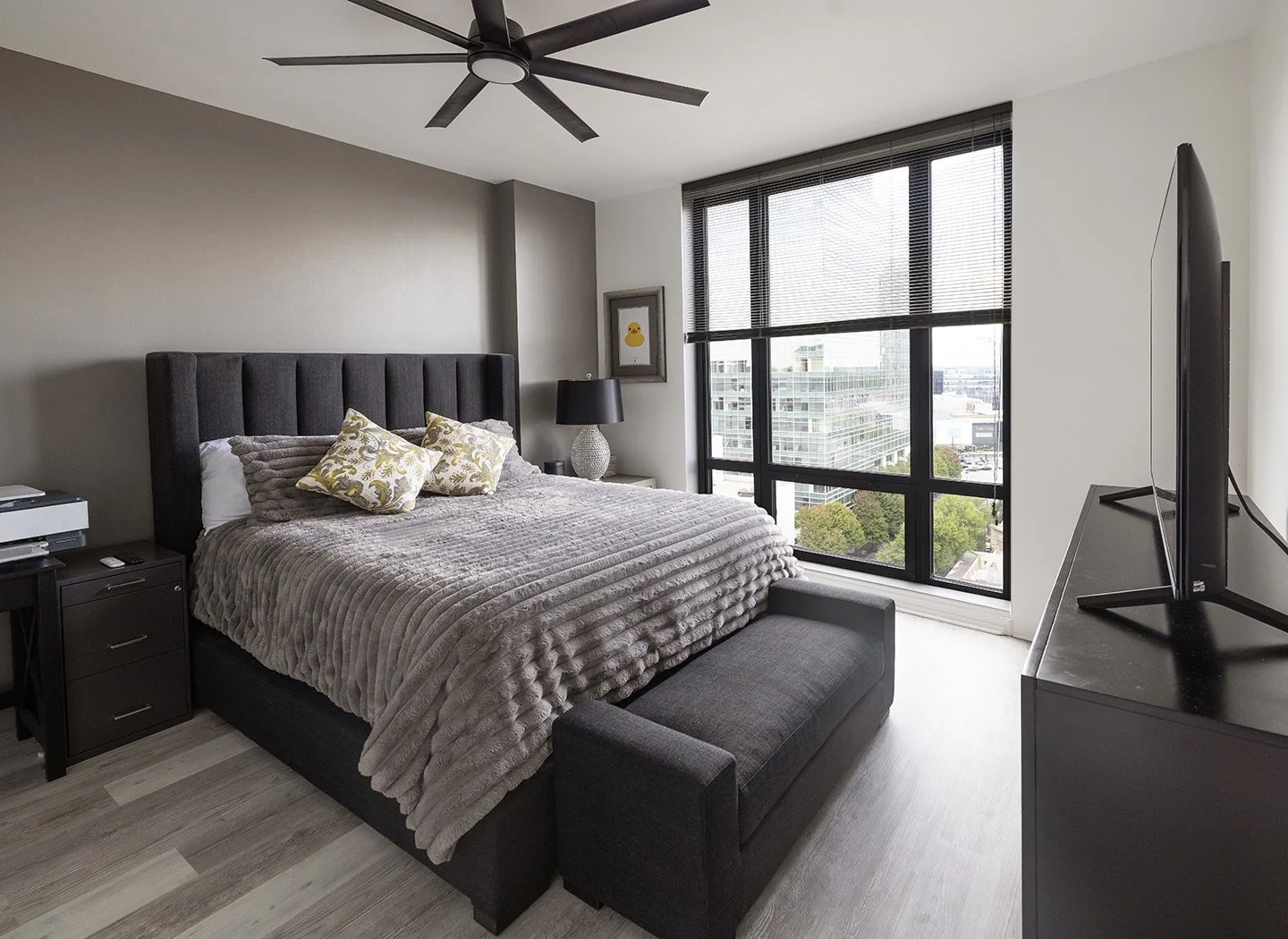 Modern bedroom with a large bed, pillows, and a bench at the foot of the bed. Black nightstand with a lamp and a framed picture on the wall. Large window with blinds revealing city buildings outside. High-rise condominium in Atlanta.
