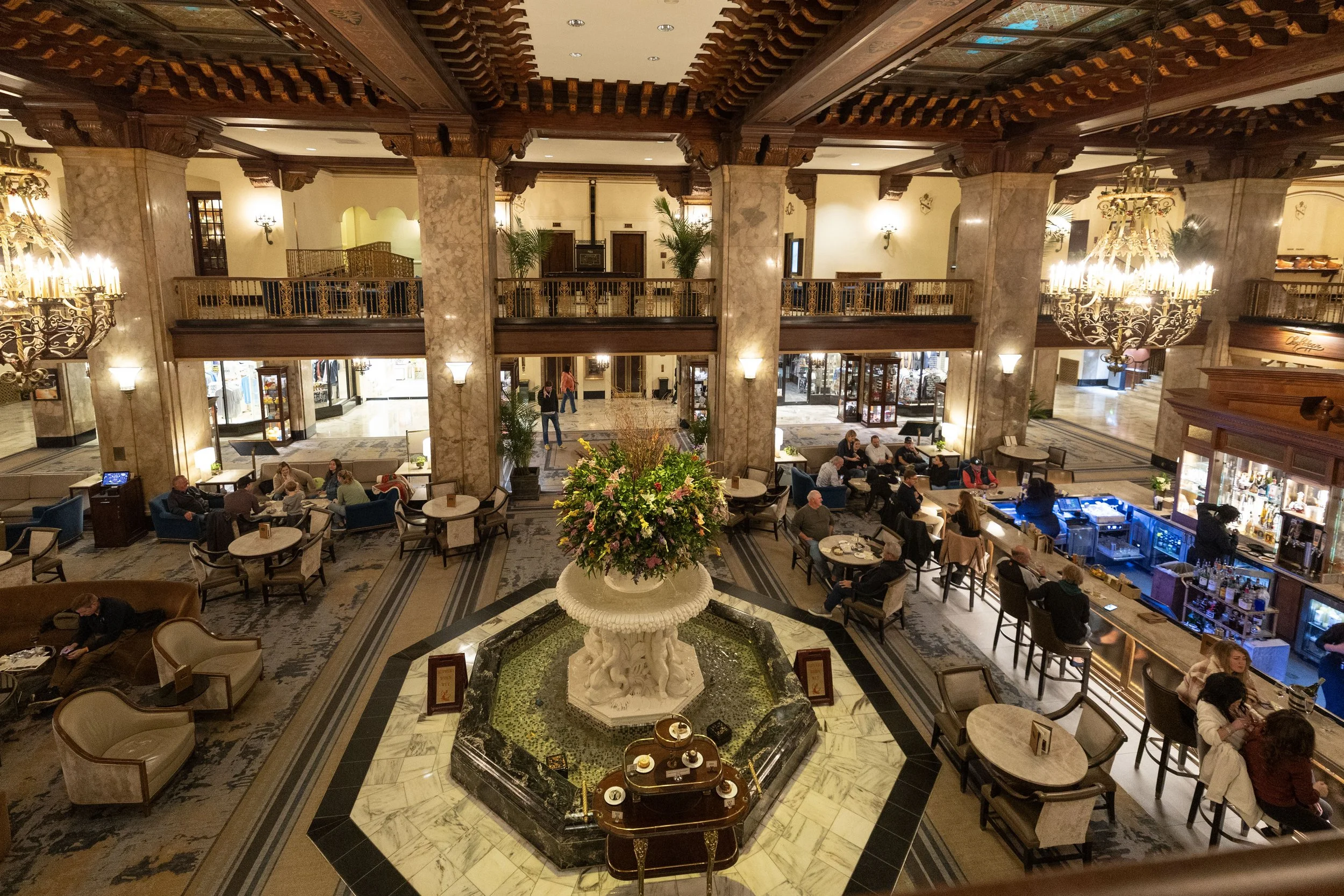 Luxurious hotel lobby with a marble fountain, large floral arrangement, seating areas, chandeliers, and a bar with patrons. Peabody Hotel Memphis.