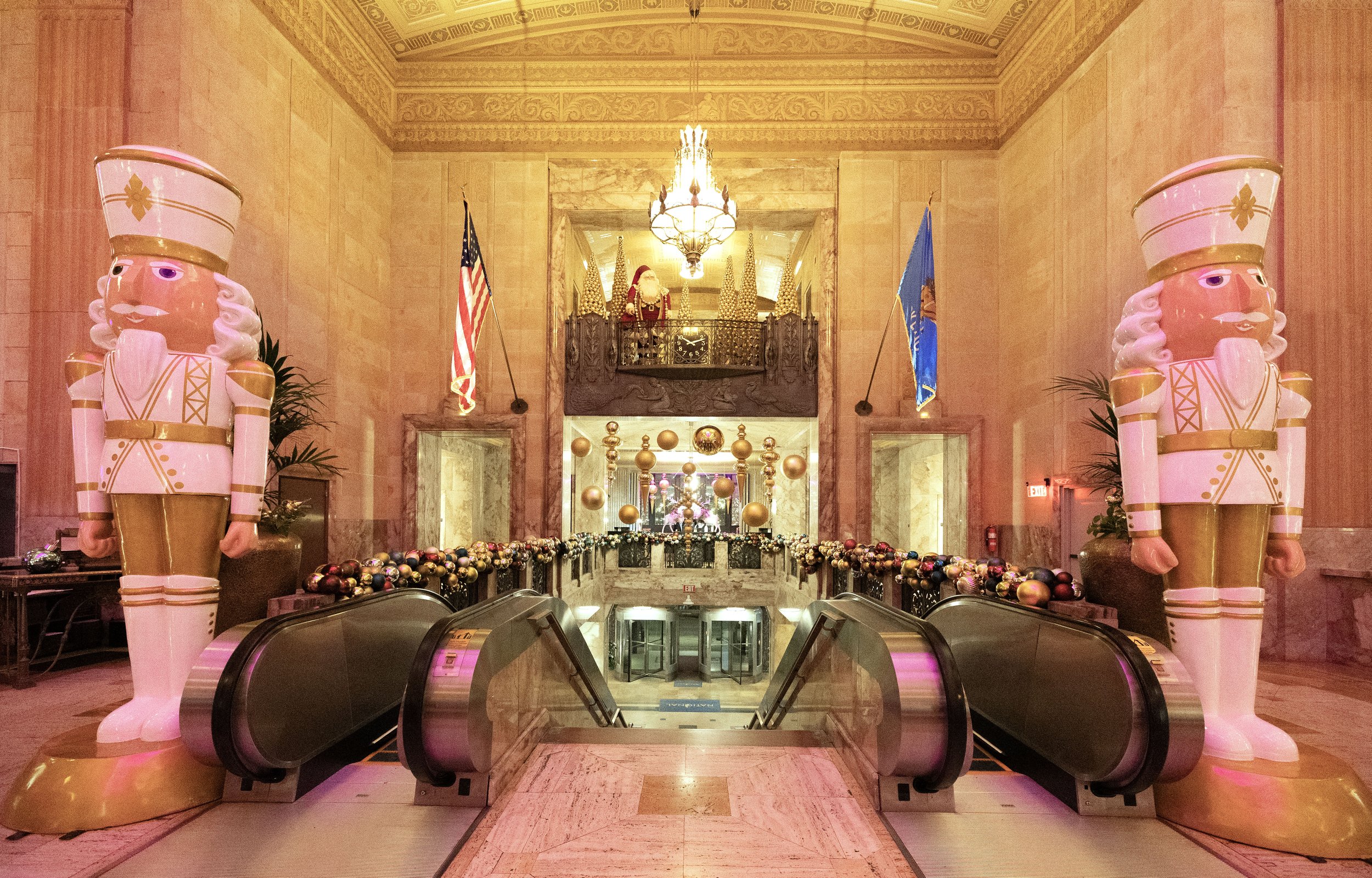 A grand hall decorated for Christmas with two large toy soldier statues on either side of an escalator. The hall features pink marble walls, flags, Christmas ornaments, and a Santa Claus figure on an upper balcony. The National Hotel, Oklahoma City.