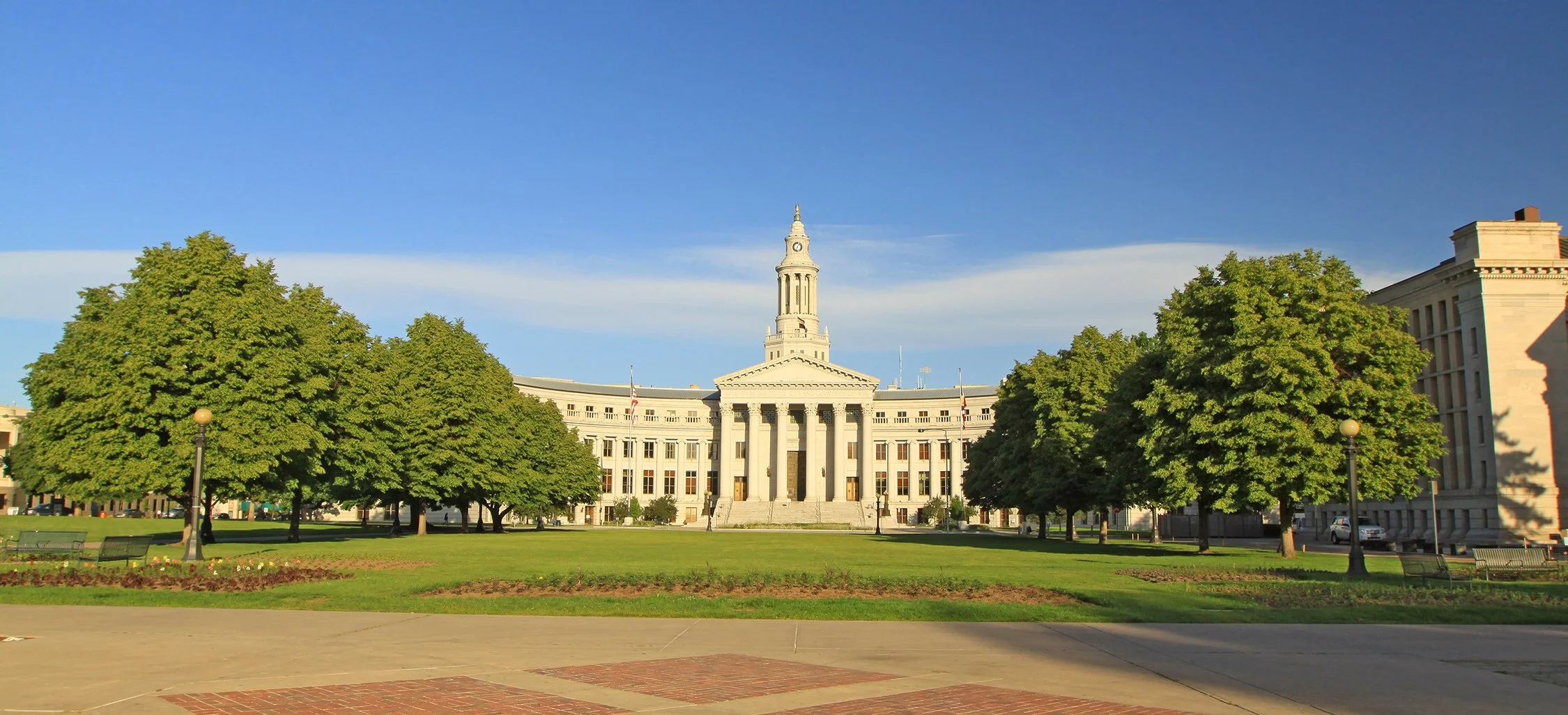 A government building with a tall clock tower, surrounded by green trees and a park in the foreground. State capitol Denver.