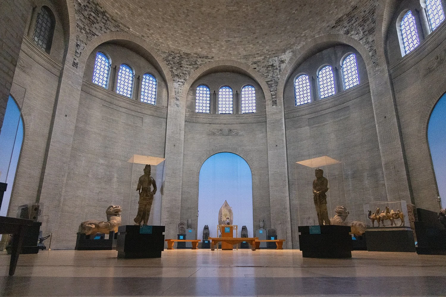 Interior of a museum with statues and artifacts displayed on black pedestals, high arched windows, and a focus on central artifact enclosure. Penn Museum Philadelphia.