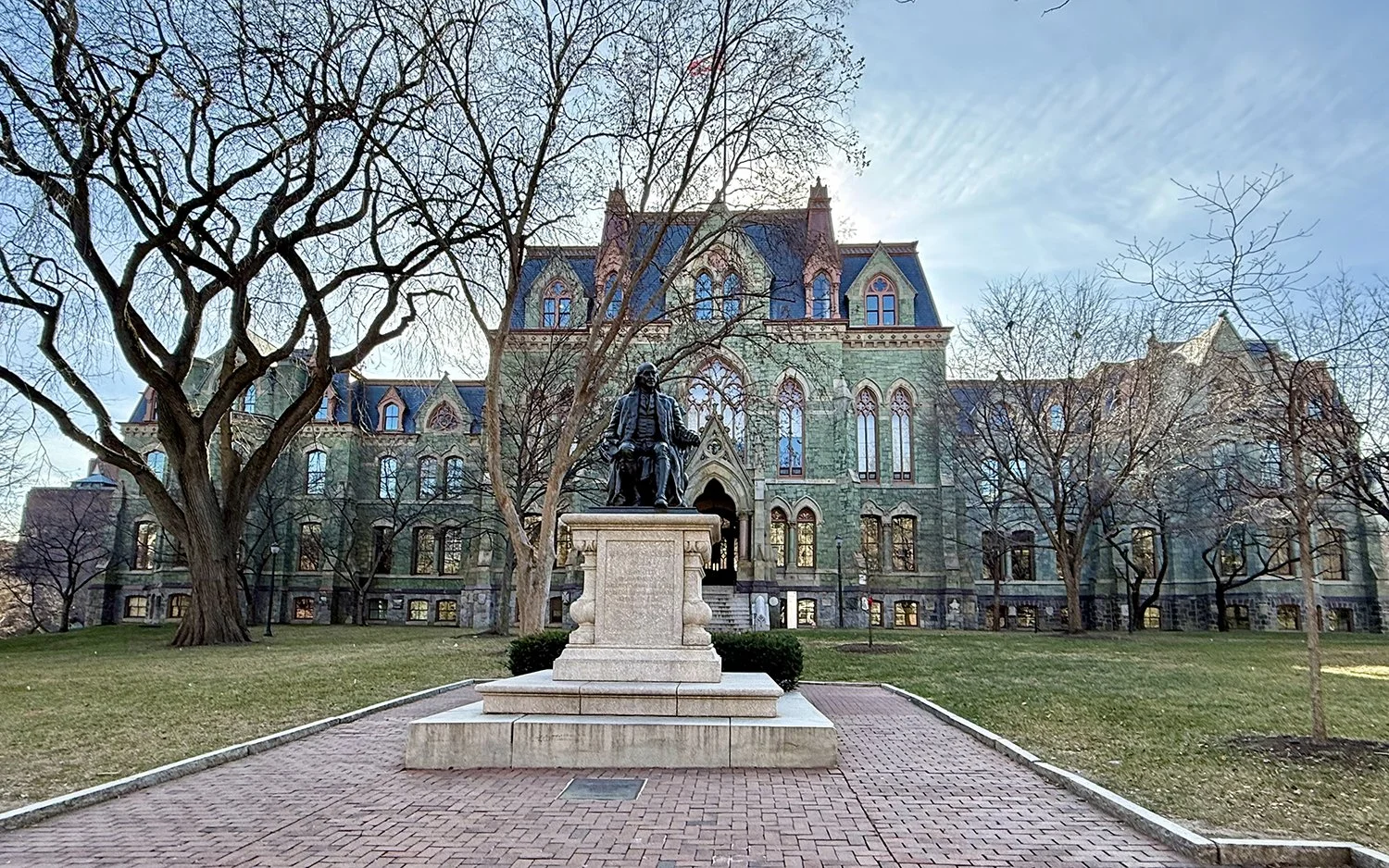 Statue of a seated man on a stone pedestal in front of a large, historic Gothic-style building with tall windows and a brick walkway. University of Pennsylvania campus building.