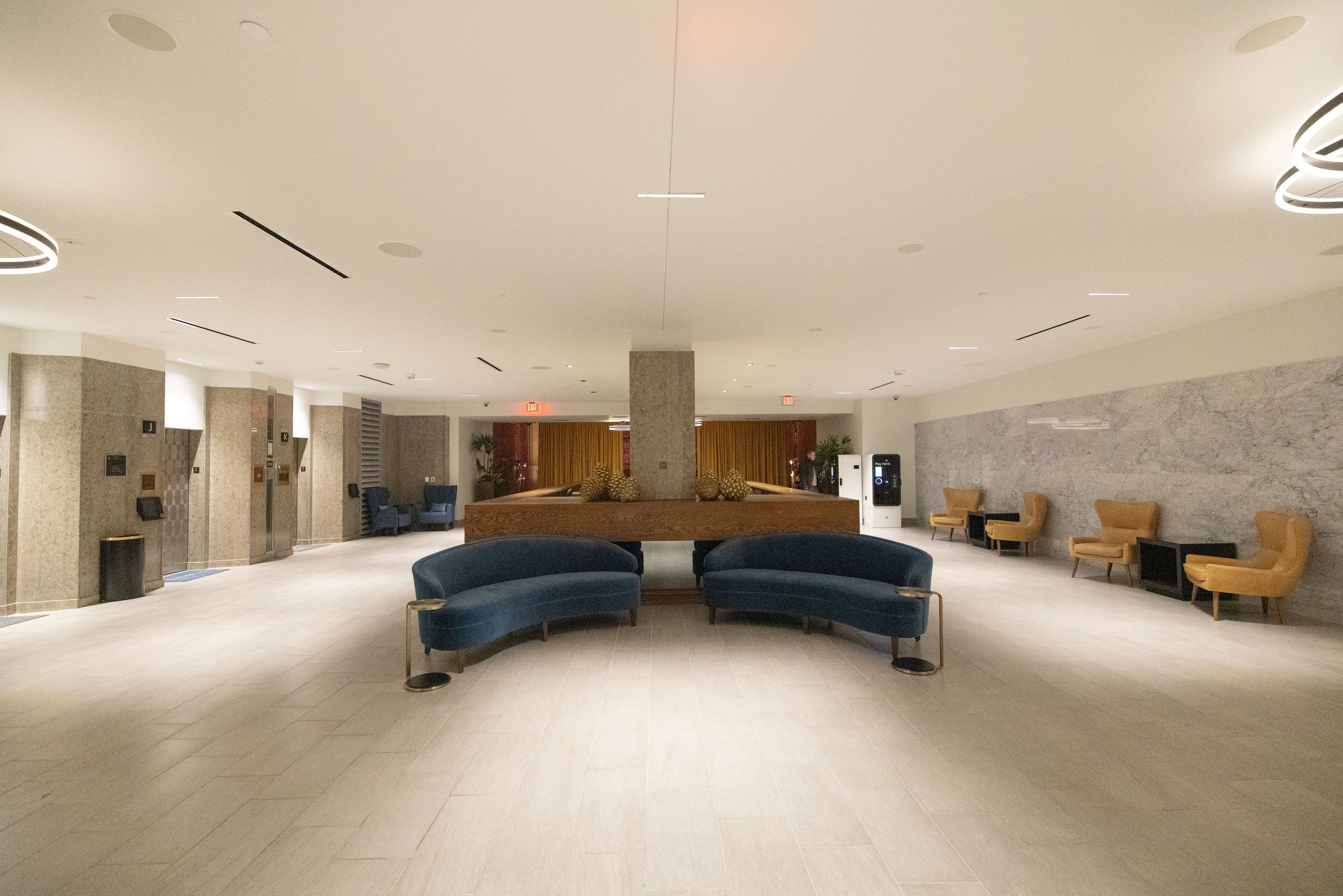 A spacious, modern hotel lobby with beige tiled flooring, a white ceiling with recessed lighting, and a central seating area with two curved dark blue velvet sofas.  The National Hotel, Oklahoma City.