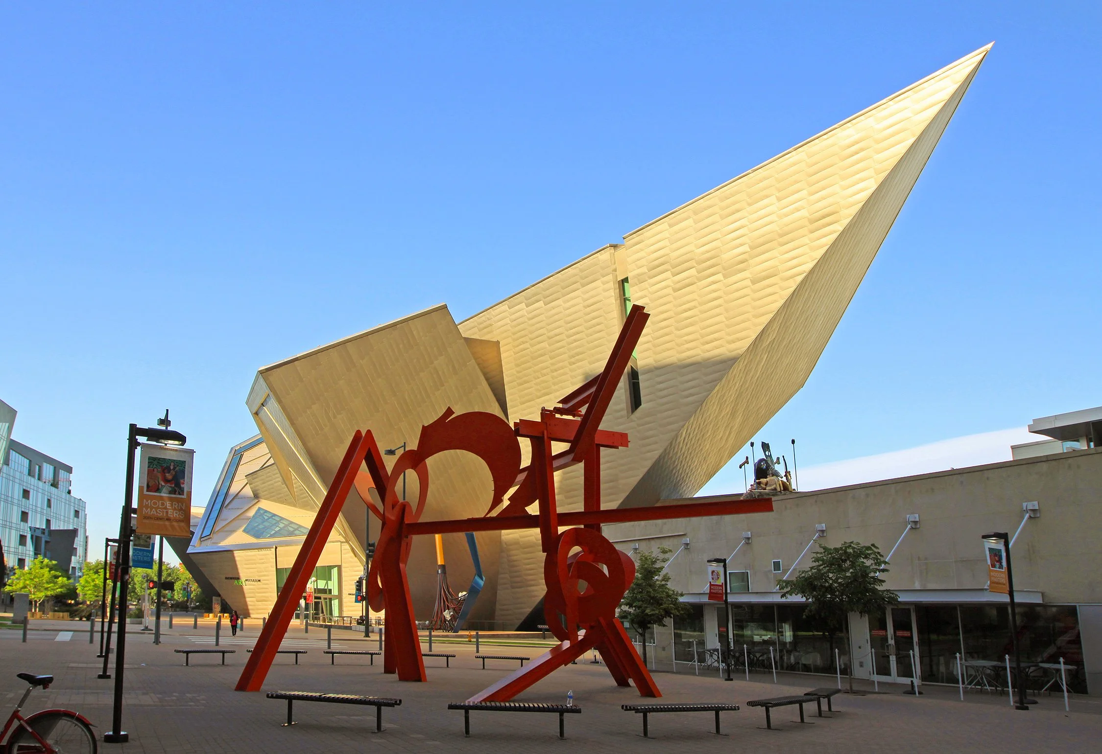 Modern building with angular gold walls, a large red abstract sculpture in the foreground, benches, trees, and banners in an outdoor plaza. Denver Art Museum.