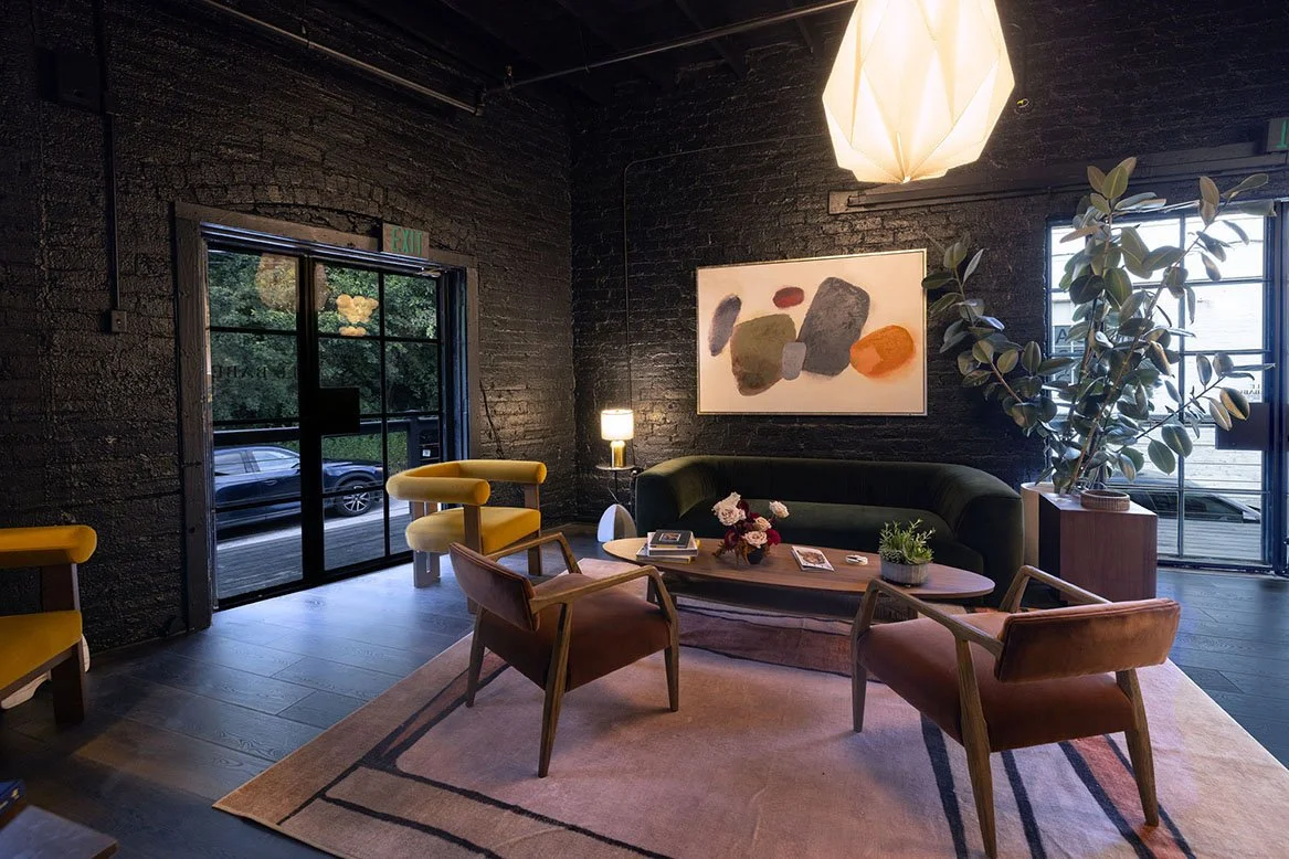 Modern interior living room with a black brick wall, large windows, a dark green sofa, four chairs, a wooden coffee table with books and flowers, a tall potted plant, abstract artwork, a ceiling lamp, and a rug. Le Babe Atlanta.