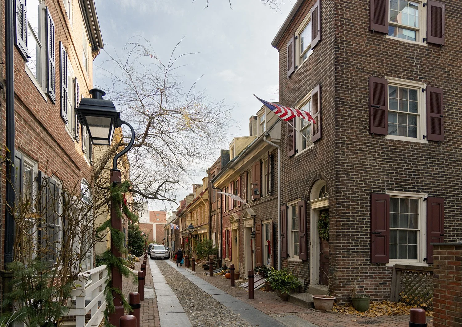 A narrow brick-lined street in a historic neighborhood decorated for Christmas. The buildings are made of brick with window shutters. Colonial street in Philadelphia.