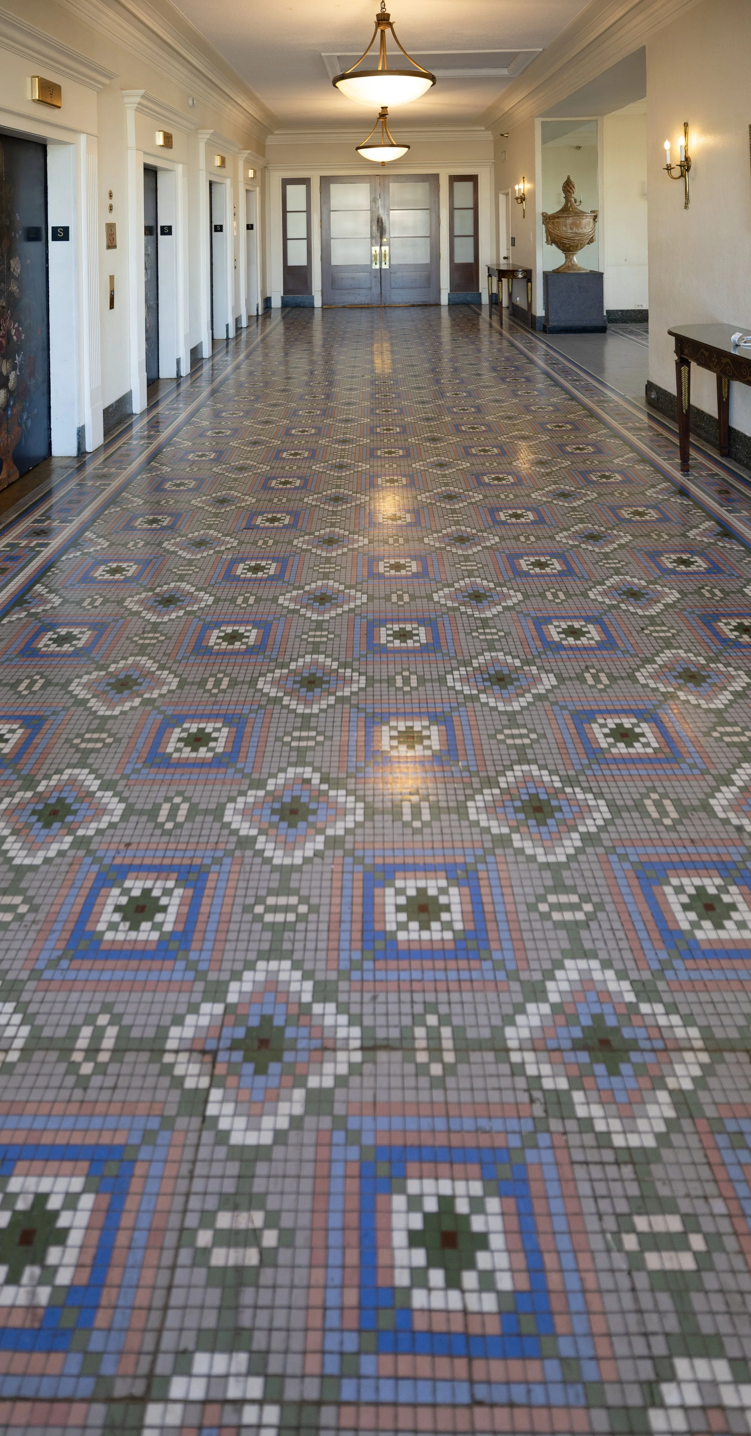 Elegant hallway with patterned tile floor, white walls, and antique decor, including a large urn on a pedestal, wall sconces, and ceiling lights. Peabody Hotel Memphis.
